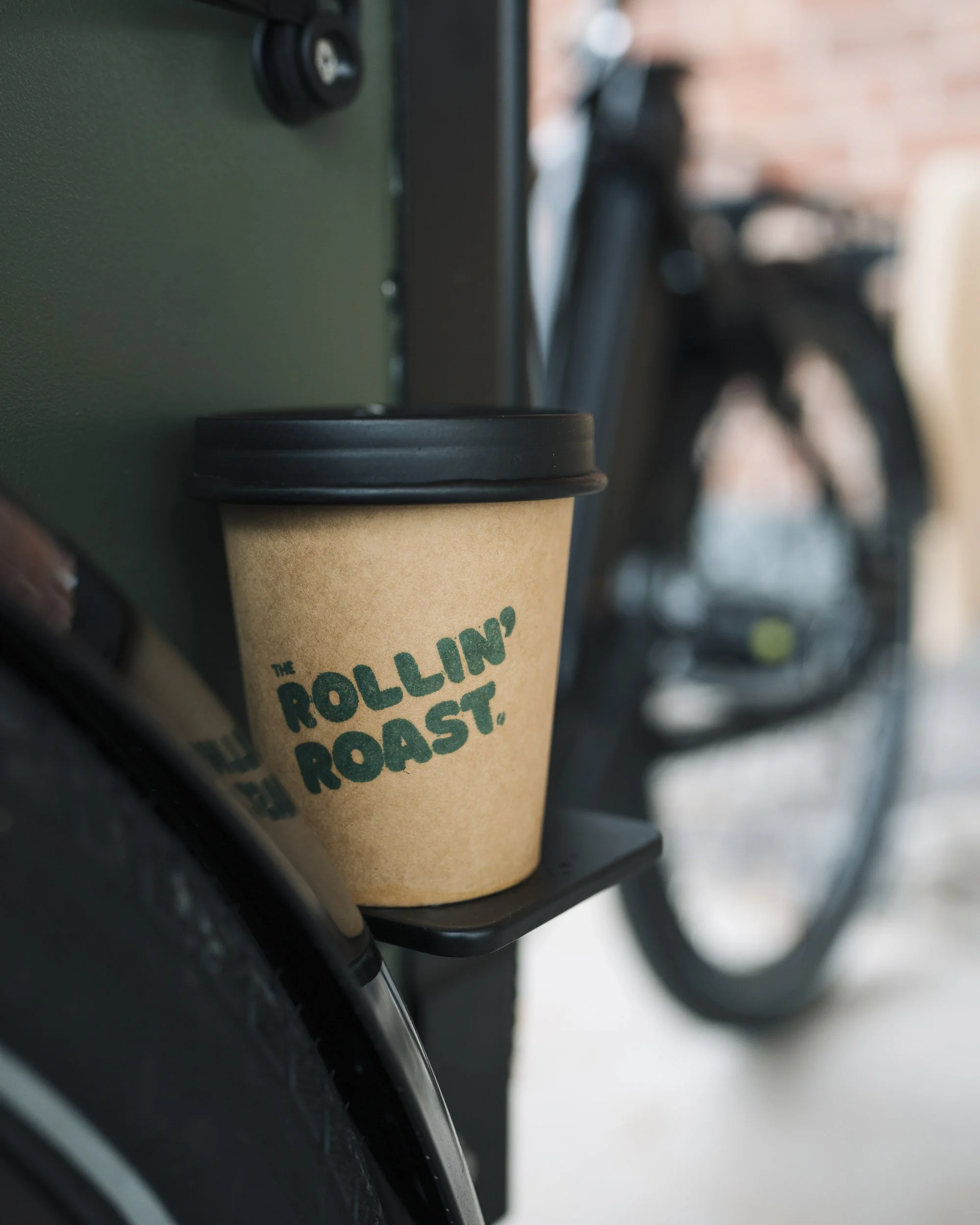 Close-up of a disposable coffee cup with a black lid mounted on a bike. The cup has a logo that reads 'The Rolling Coast' in green text. The background shows blurred tires and a brick wall.