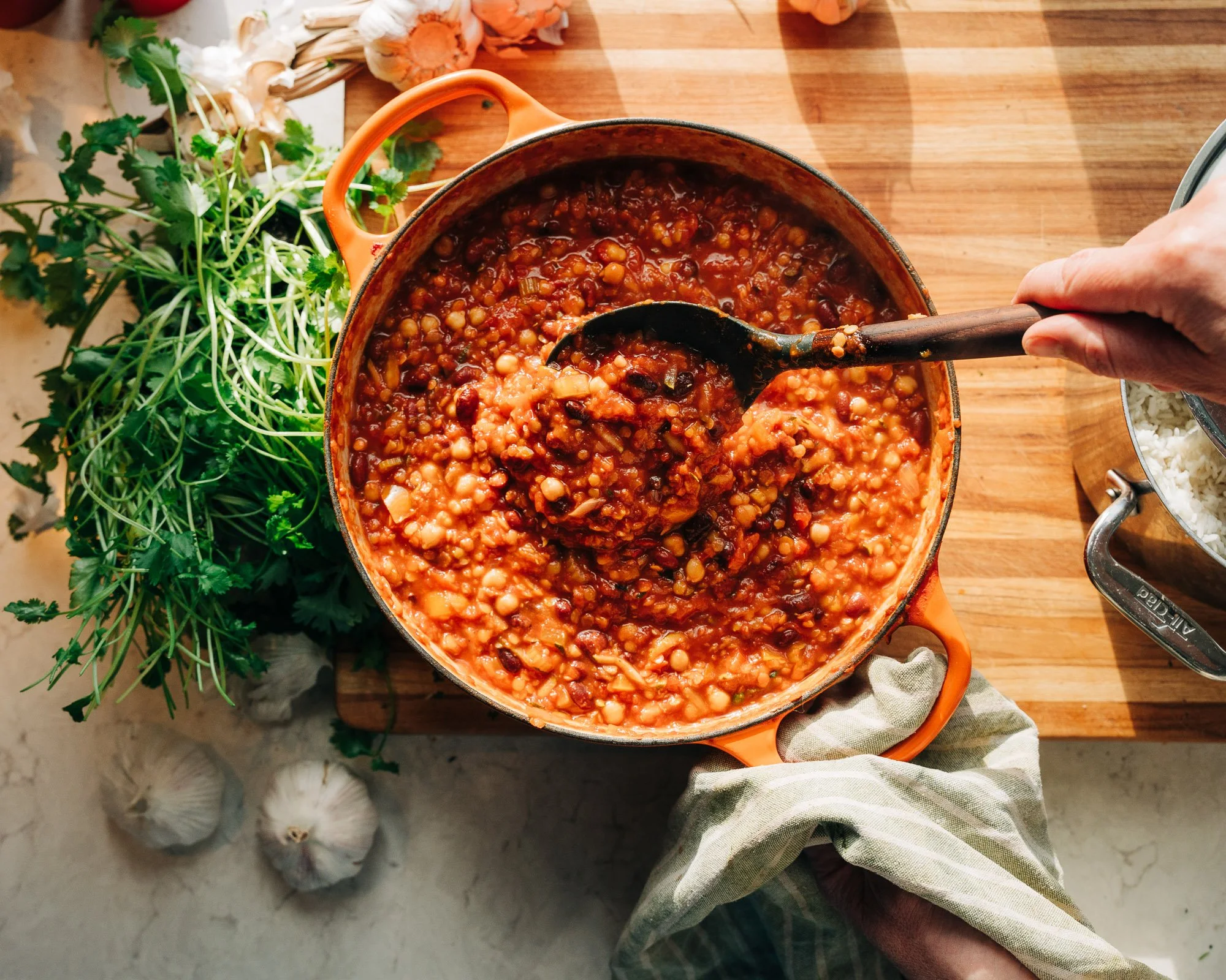 A person stirring a pot of cooked chili with beans on a wooden countertop. Whole garlic bulbs and fresh herbs are around the pot, and the person's hand is holding a spoon.