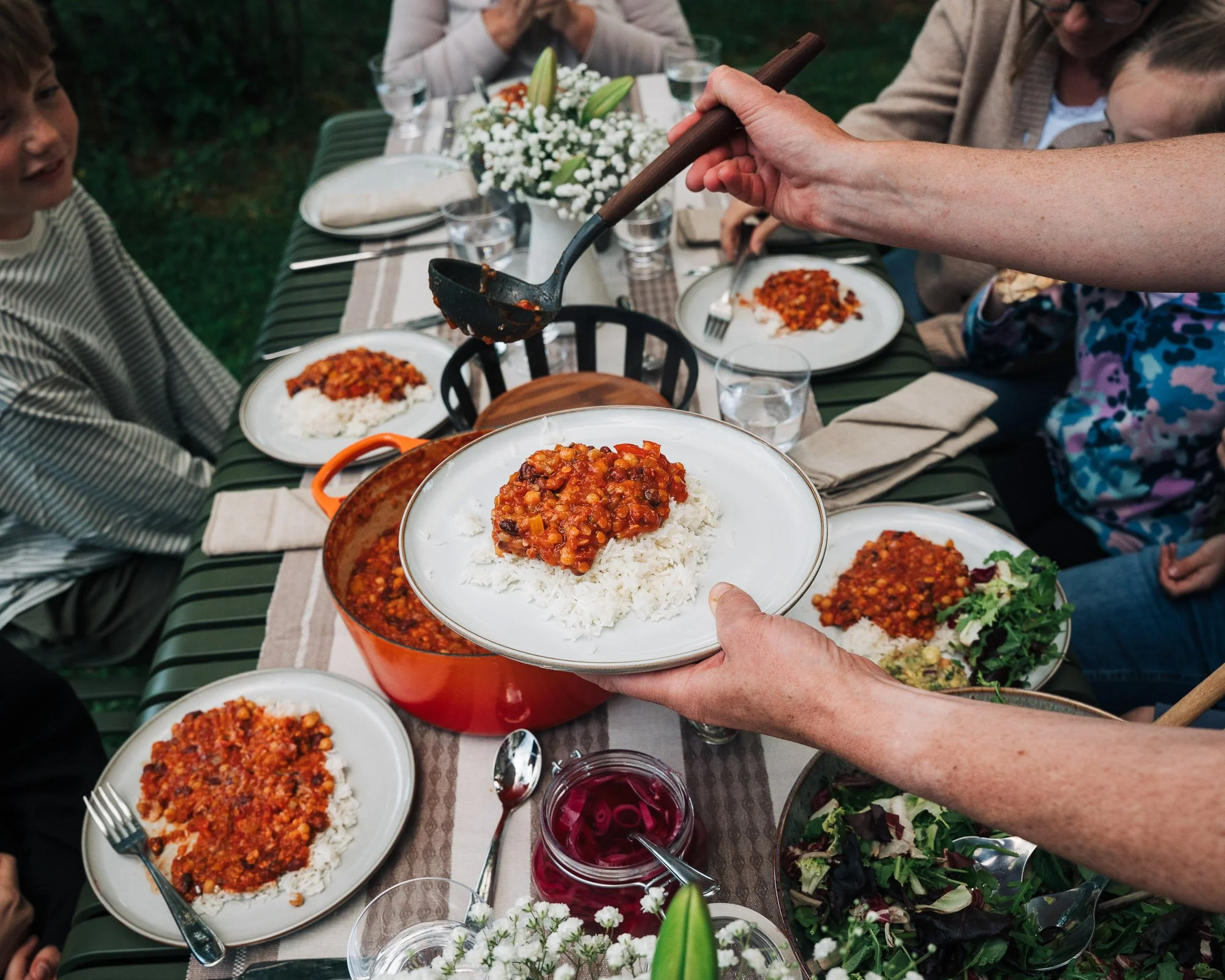 People at an outdoor dinner table sharing plates of rice with chili and vegetables, with floral centerpieces and glasses of drinks.