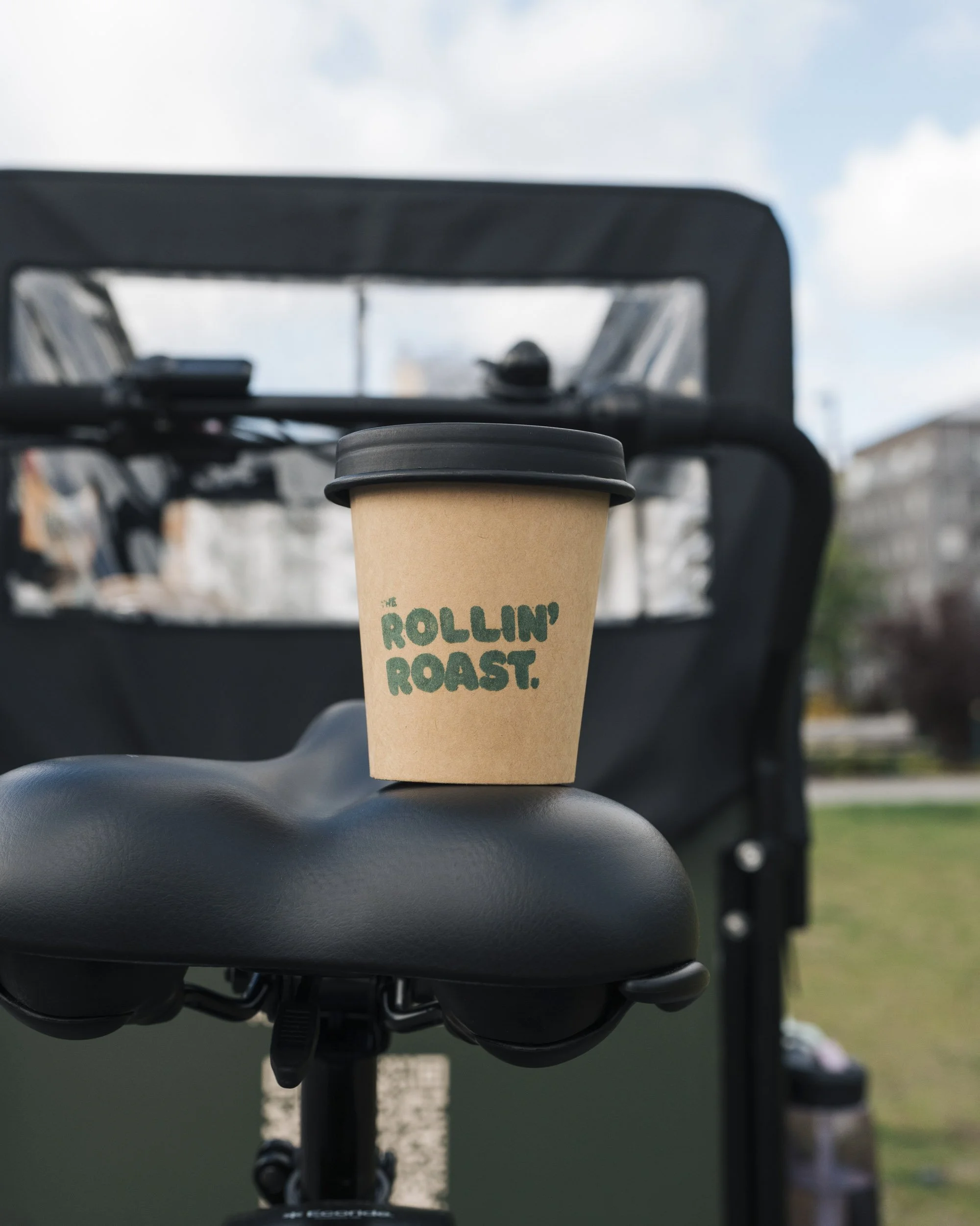 Close-up of a disposable coffee cup with a black lid placed on the black seat of a bicycle or motorcycle, with a blurred background including buildings and a cloudy sky.