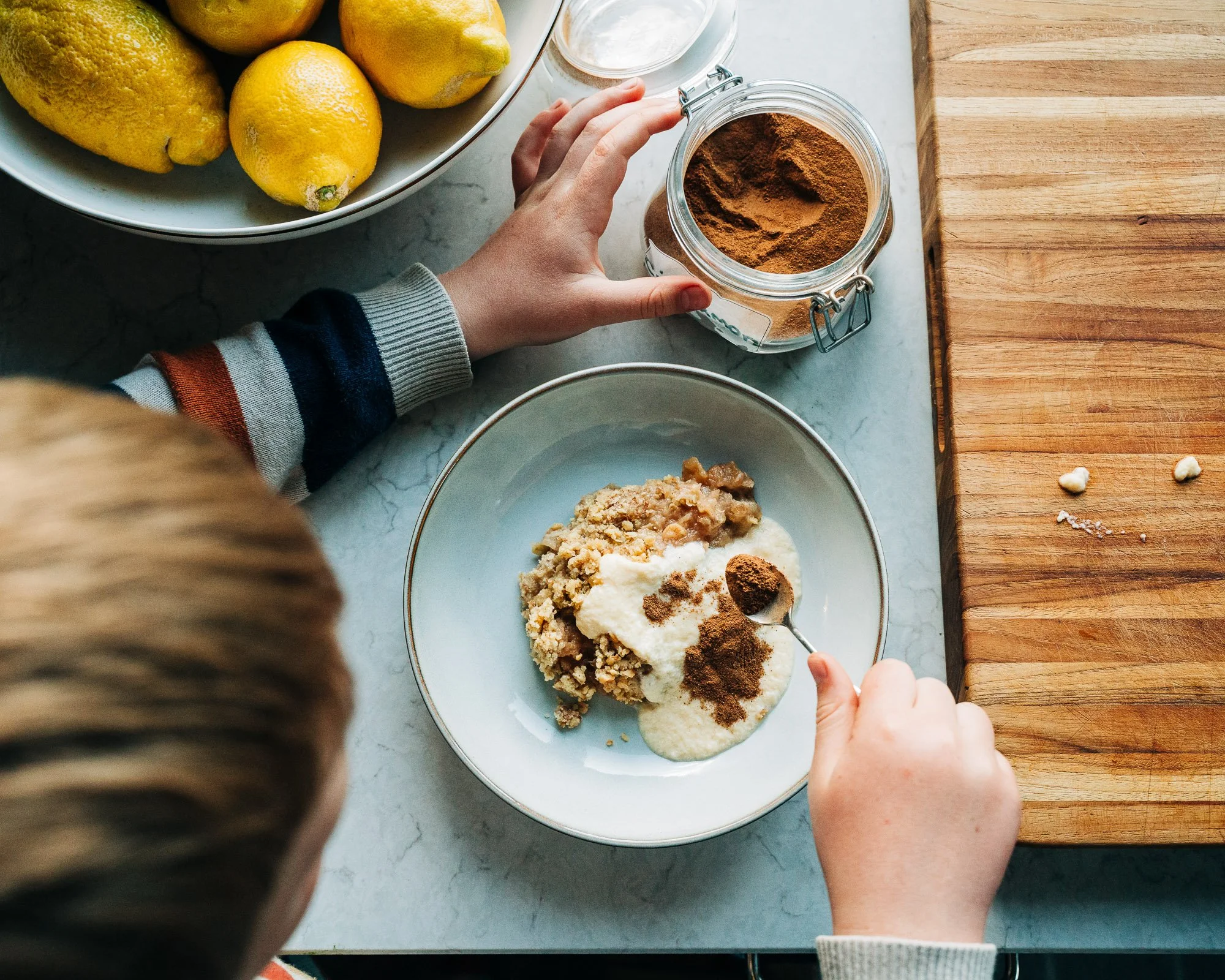 Person using a spoon to add cinnamon to a dessert in a white bowl on a marble countertop, with lemons in a bowl and a wooden cutting board nearby.