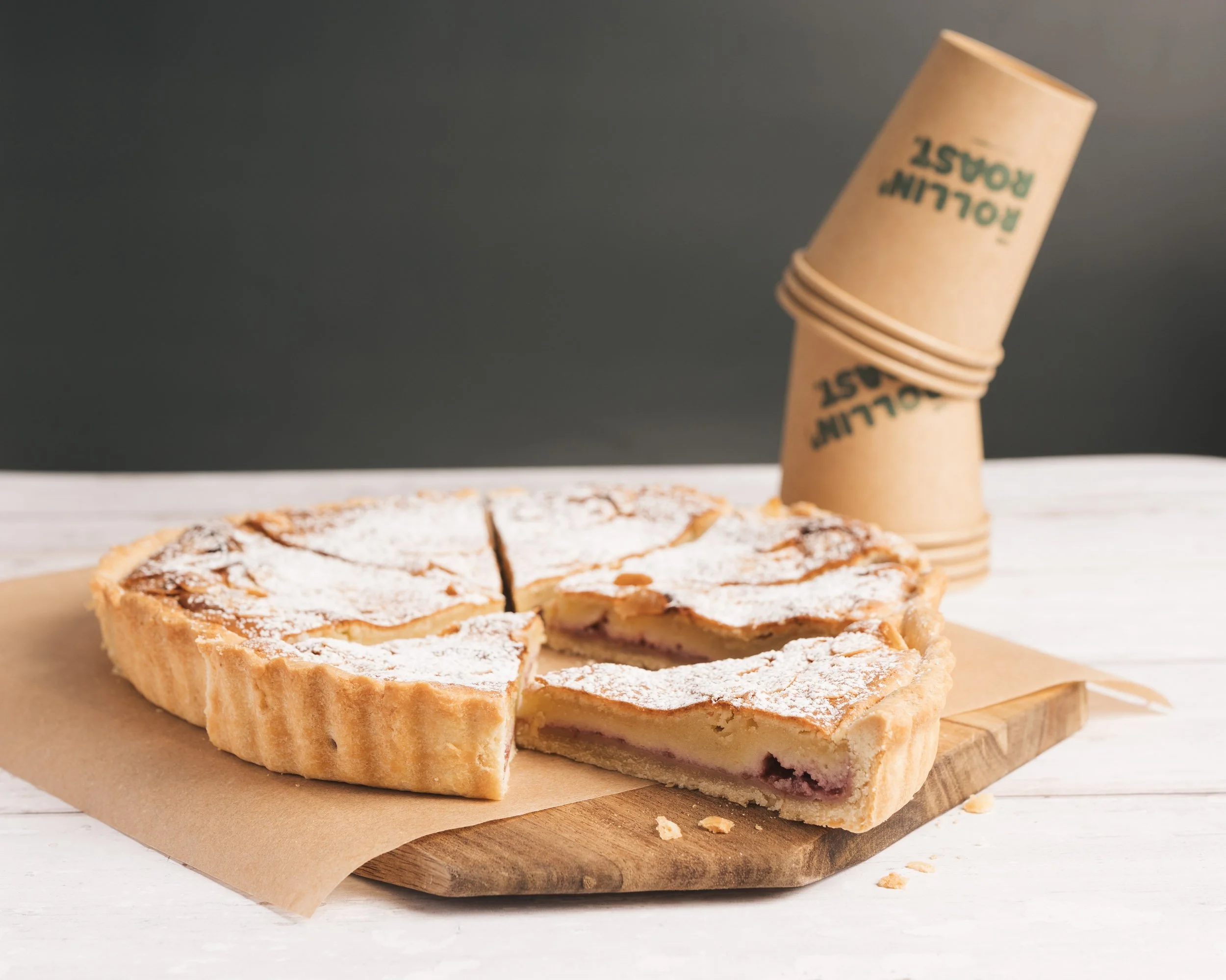Sliced pastry with powdered sugar on top, placed on a wooden cutting board, with a group of stacked paper cups in the background.