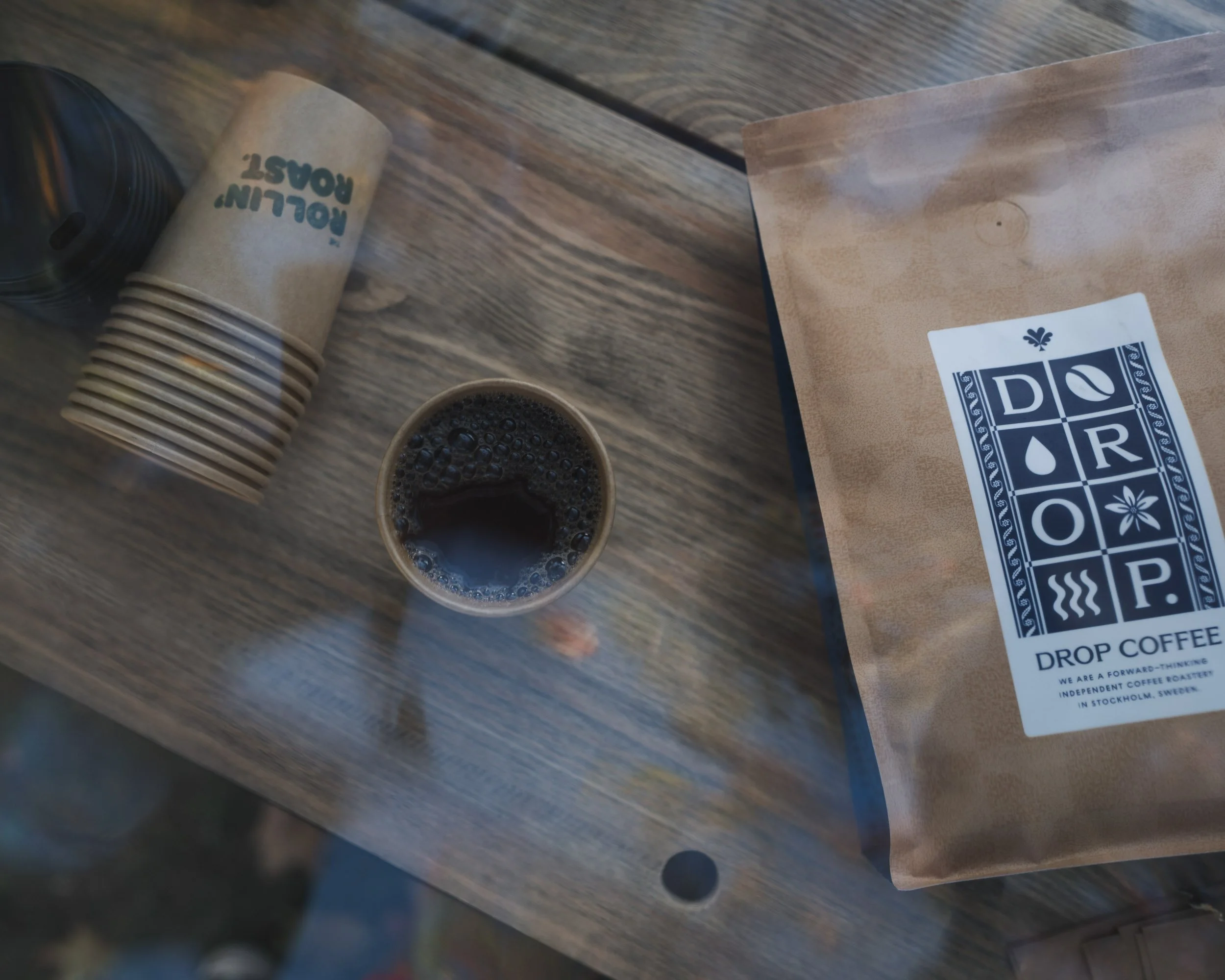 A bag of coffee beans, a cup of brewed coffee, a paper coffee cup sleeve, and a paper bag from Dialogue Coffee Roasters on a wooden table.