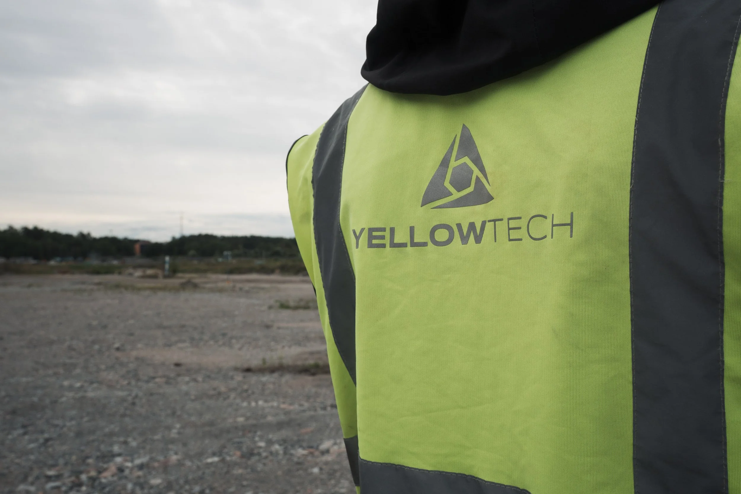 Close-up of a person wearing a bright yellow-green safety vest with the logo and name 'YELLOWTECH' on the front, standing outdoors on a cloudy day with a landscape of gravel and distant trees.