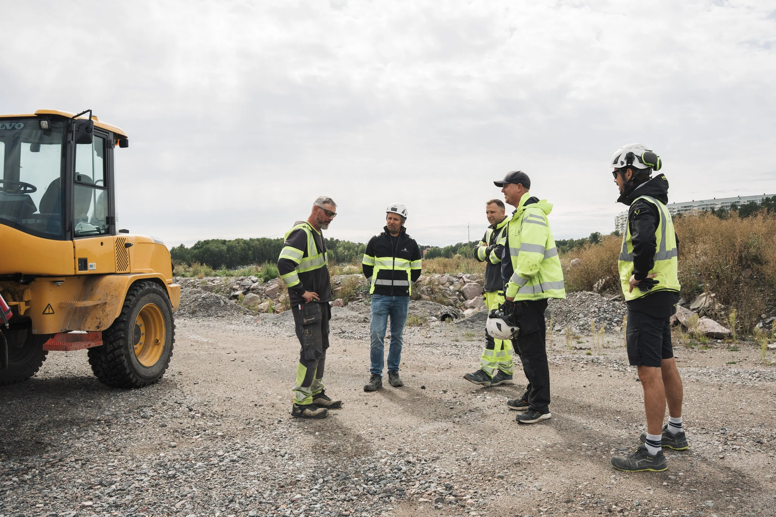 Group of five men in safety gear having a discussion at a construction site with a yellow construction vehicle nearby, in an outdoor area with rocky ground and vegetation in the background.
