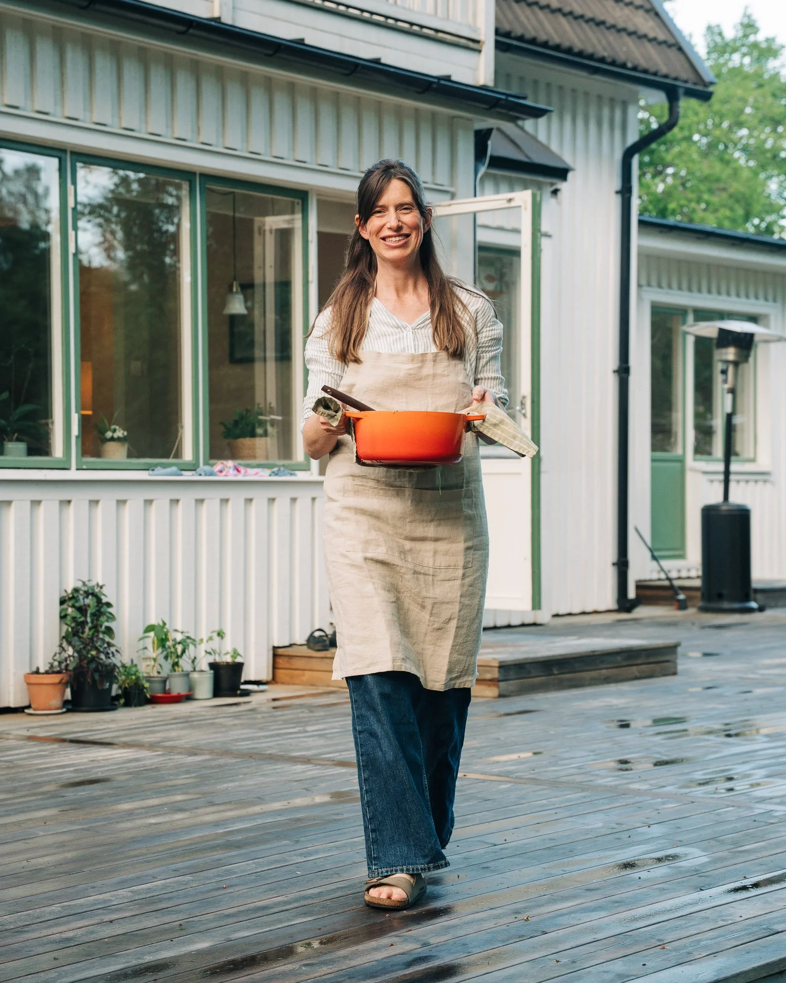 A woman smiling and holding a red pot while walking on a wooden deck outside a house with large windows and potted plants.