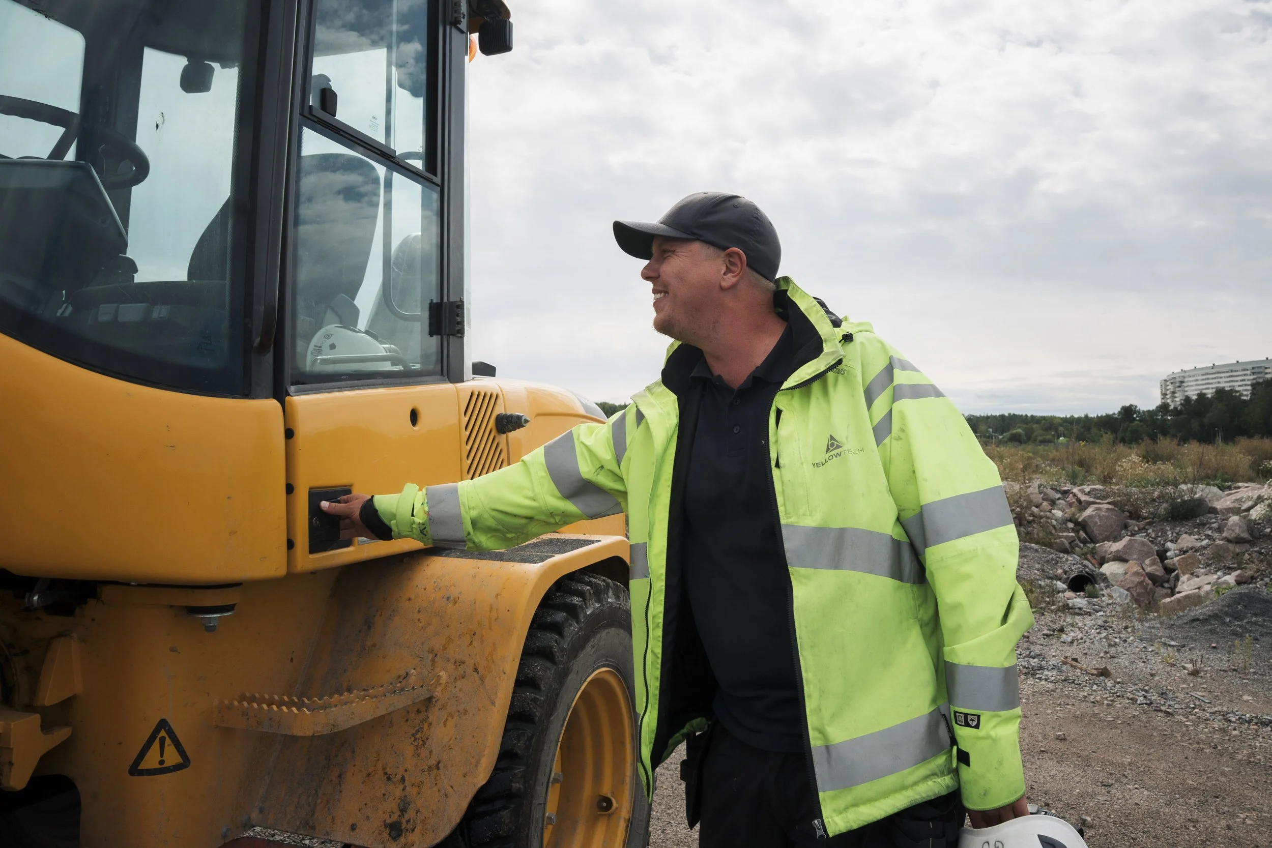 A construction worker is standing next to a yellow road roller compactor, smiling, wearing a black cap and a bright yellow-green safety jacket, and holding a white safety helmet in his right hand. The background shows a cloudy sky and a distant build