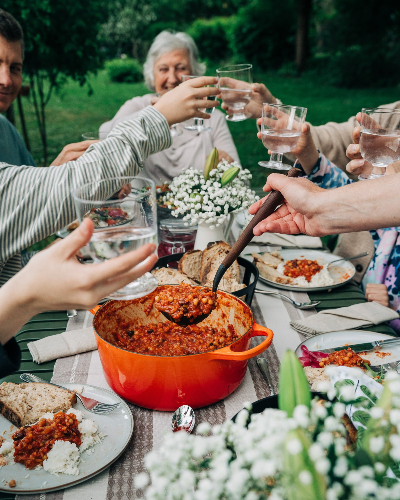 Several people raising glasses in a toast at an outdoor dinner party with a table of food including a large pot of chili, bread, and various side dishes, surrounded by greenery.
