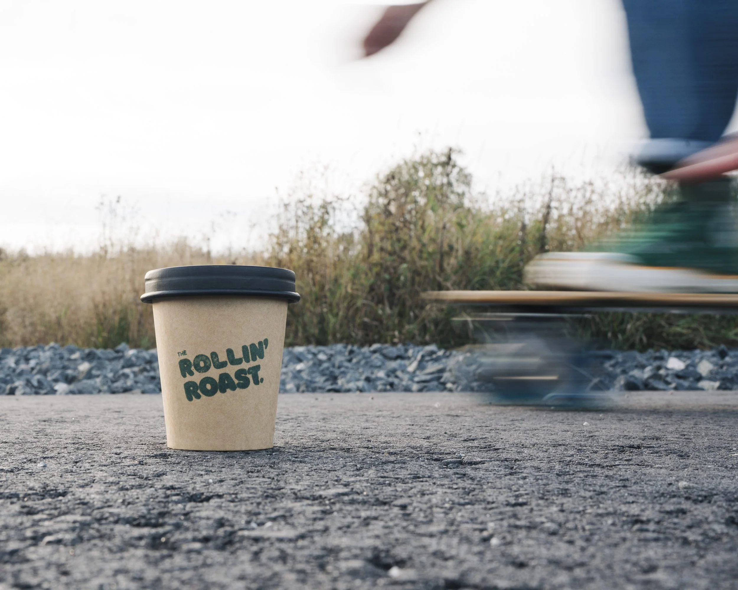 A disposable coffee cup with a black lid placed on a gravel or asphalt surface outdoors. In the background, a blurry hand is holding a tray with a person’s arm visible, and some trees and sky are also visible.