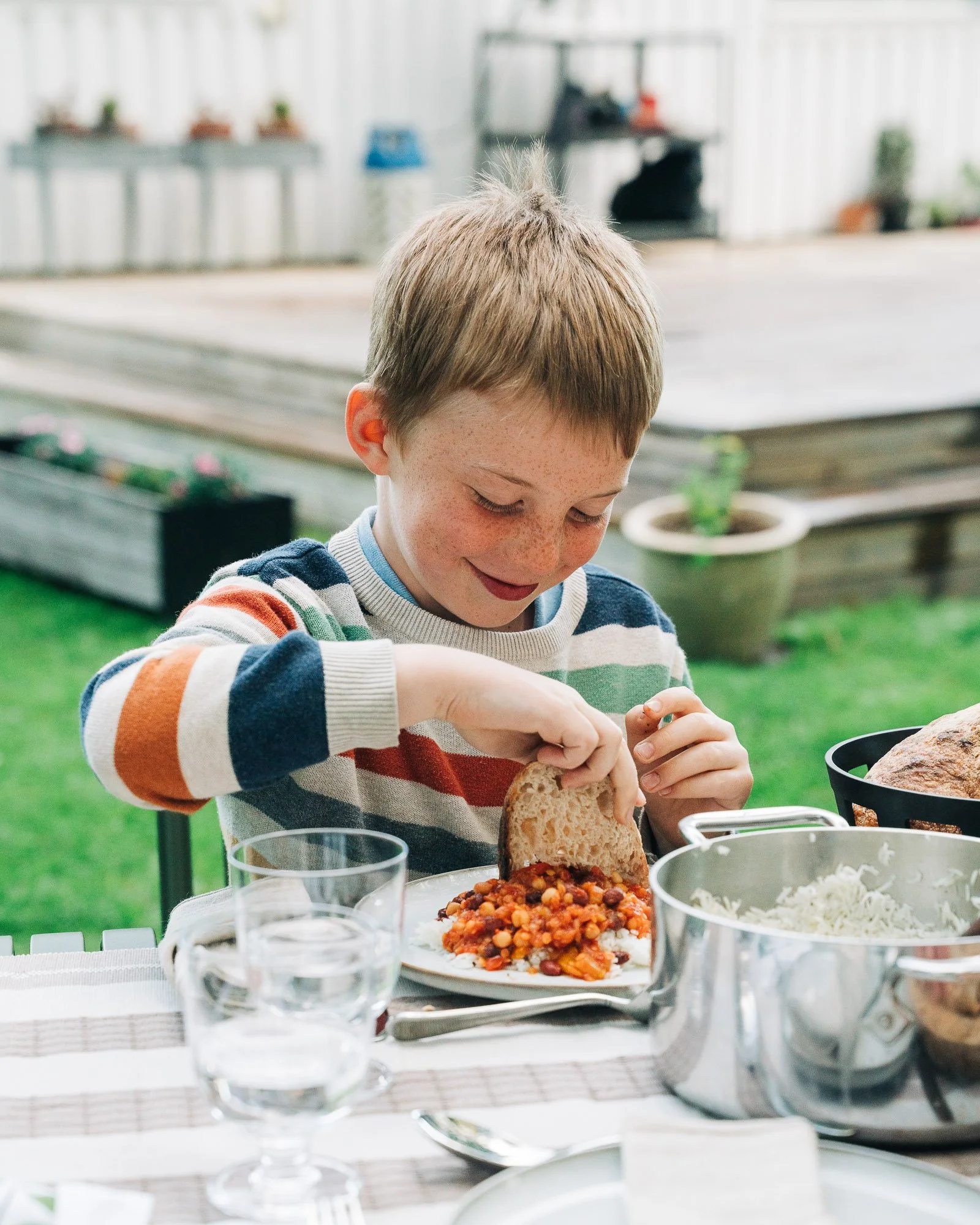 Child with light brown hair and freckles, wearing a striped sweater, smiling while preparing food at an outdoor dining table with various dishes and glasses.