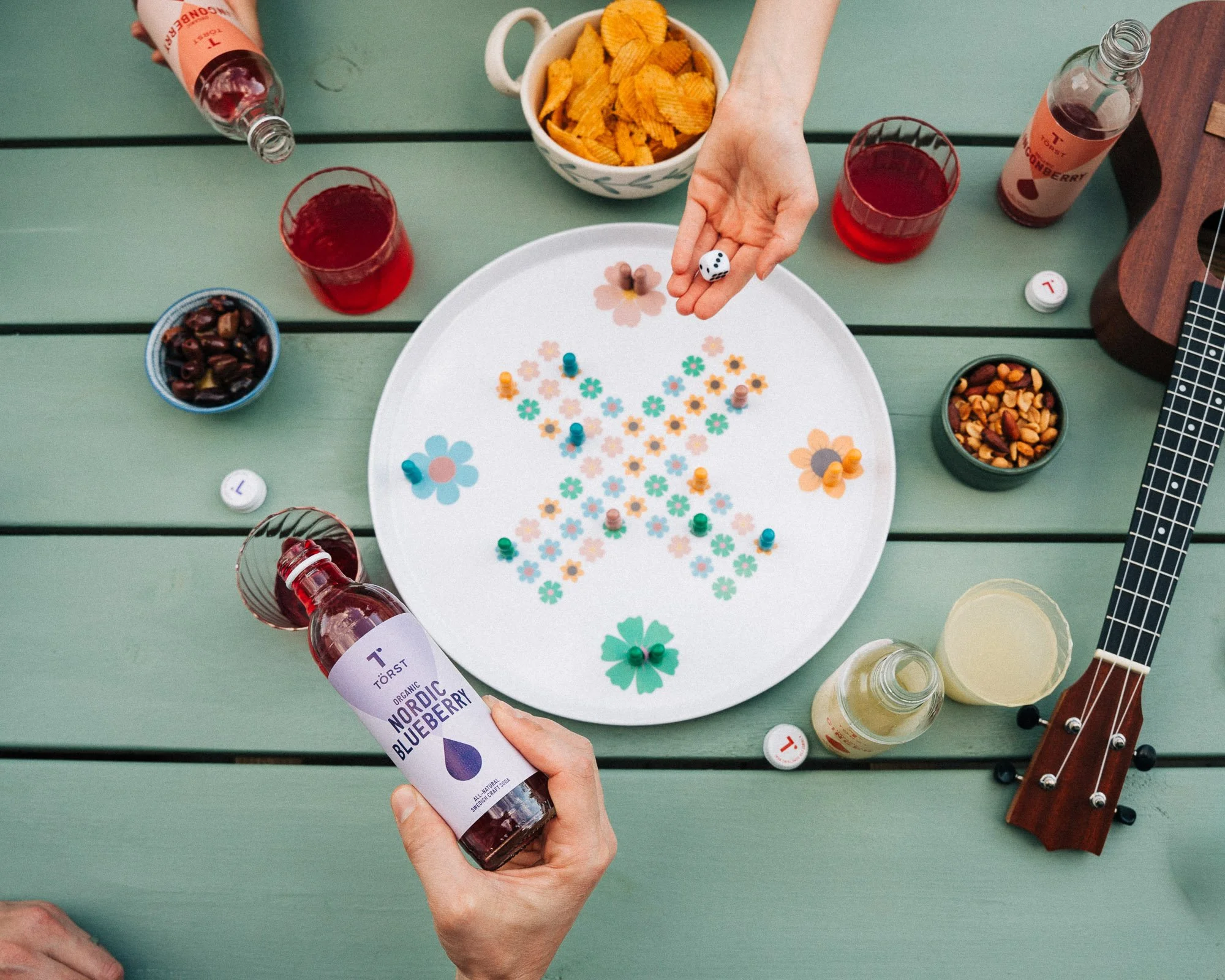 Top-down view of a person holding a bottle of Törst Nordic Organic Blueberry soda, surrounded by a green wooden table with various snacks, drinks, a guitar, and game pieces.