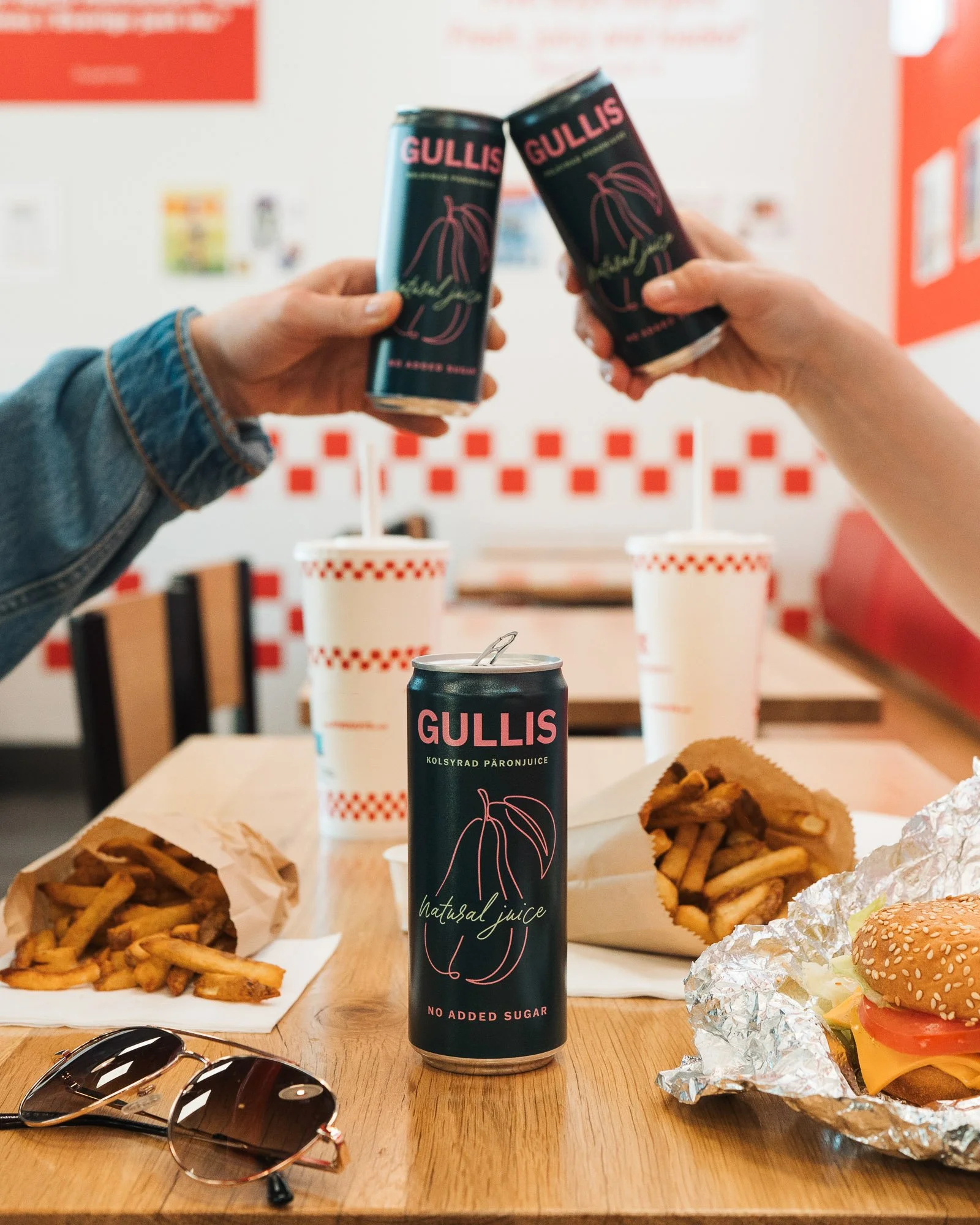 Two people holding cans of Gullis natural juice with a burger, fries, and soda on a wooden table in a fast food setting.