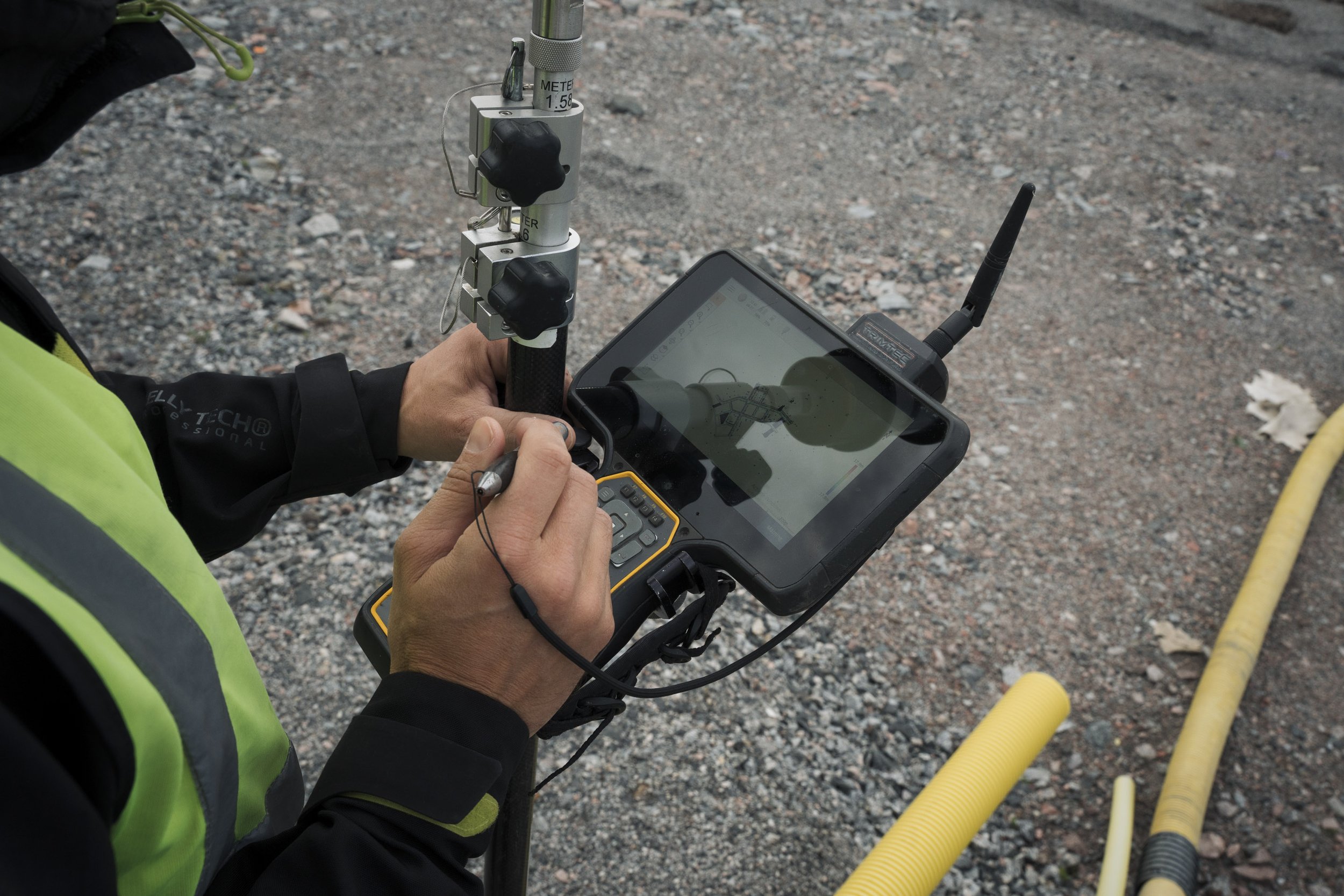 A person wearing a safety vest operating a portable measuring device with a cable connected to a tablet, on a gravel surface.