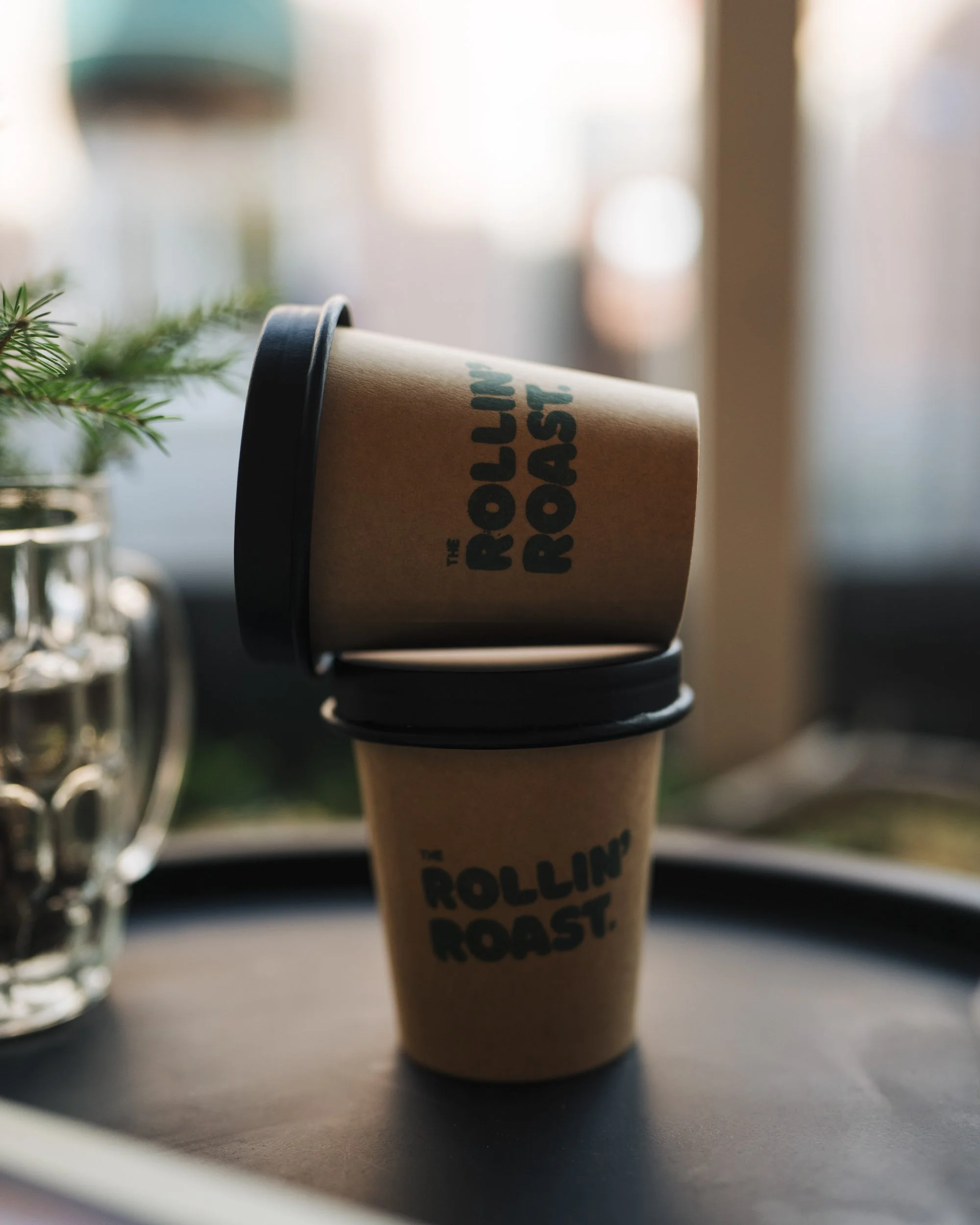A disposable coffee cup with a black lid, sitting on a dark surface near some greenery with a blurred background.