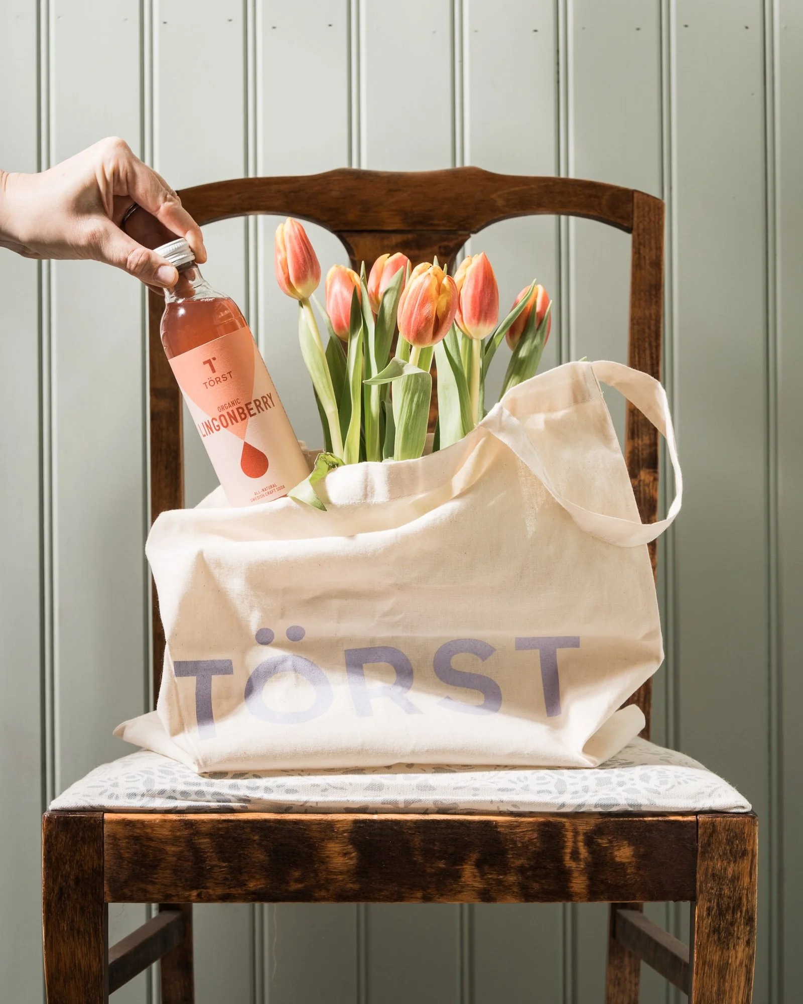 A wooden chair with a beige tote bag containing orange and yellow tulips and a bottle of Törst Lingonberry, set against a vertical green panel wall.