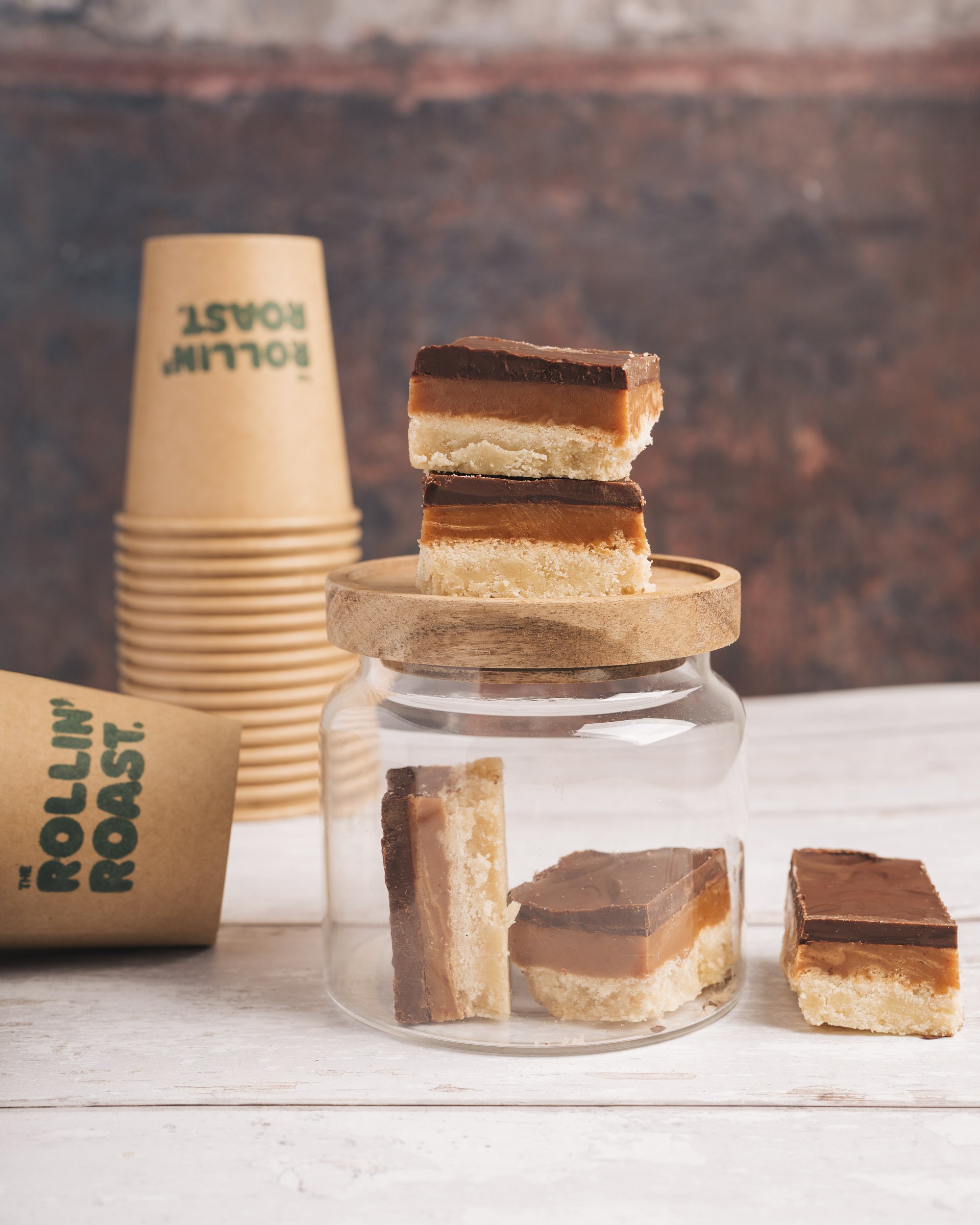 Stacked layered homemade shortbread cookies with caramel and chocolate layers on a glass cake stand, with a paper coffee cup from Folly Roast in the background.
