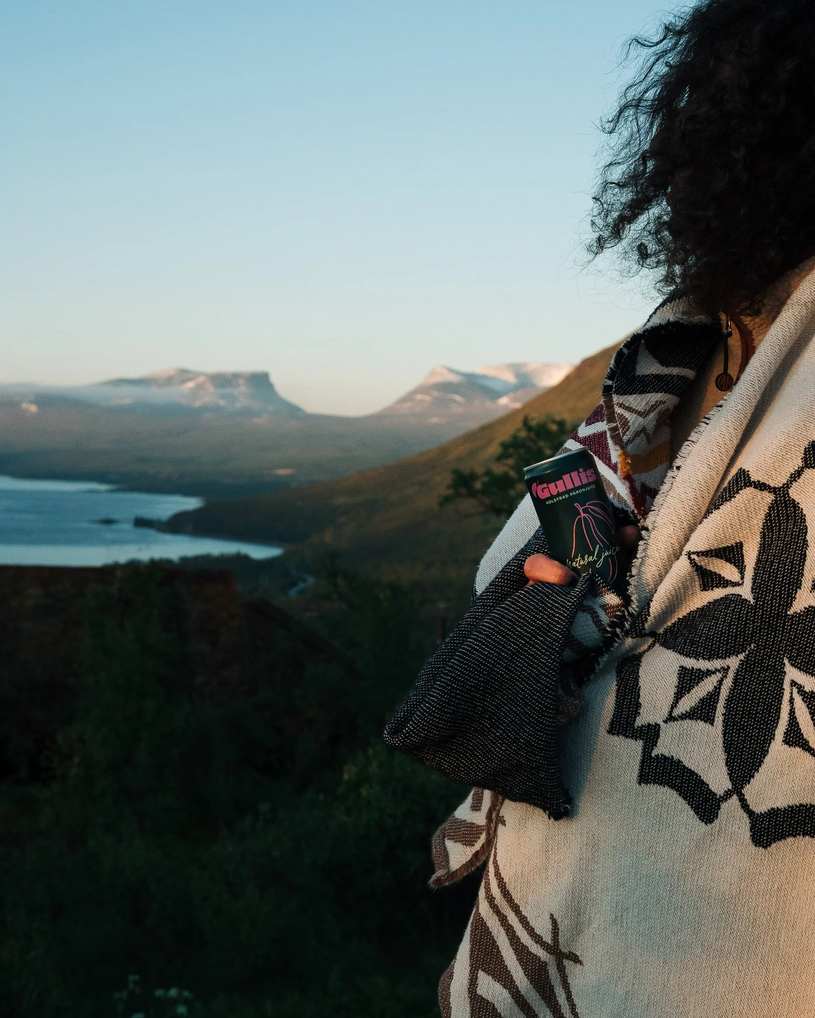 Person holding a can of juice in an outdoor landscape with mountains in the background during sunset.