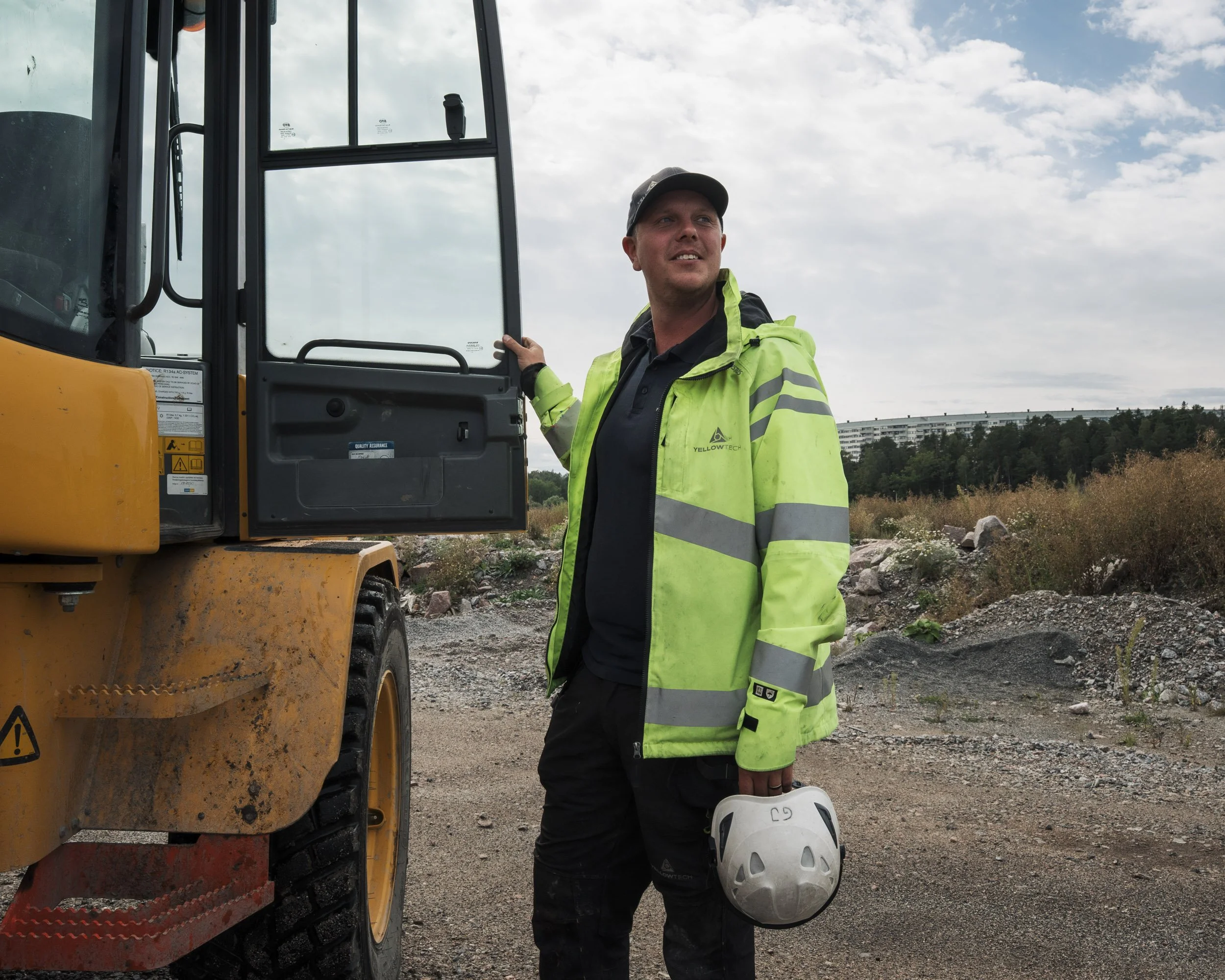A construction worker wearing a high-visibility green jacket and a baseball cap stands outdoors next to a yellow construction vehicle. The worker is holding a white safety helmet and is looking to the side with a slight smile. The background shows a 