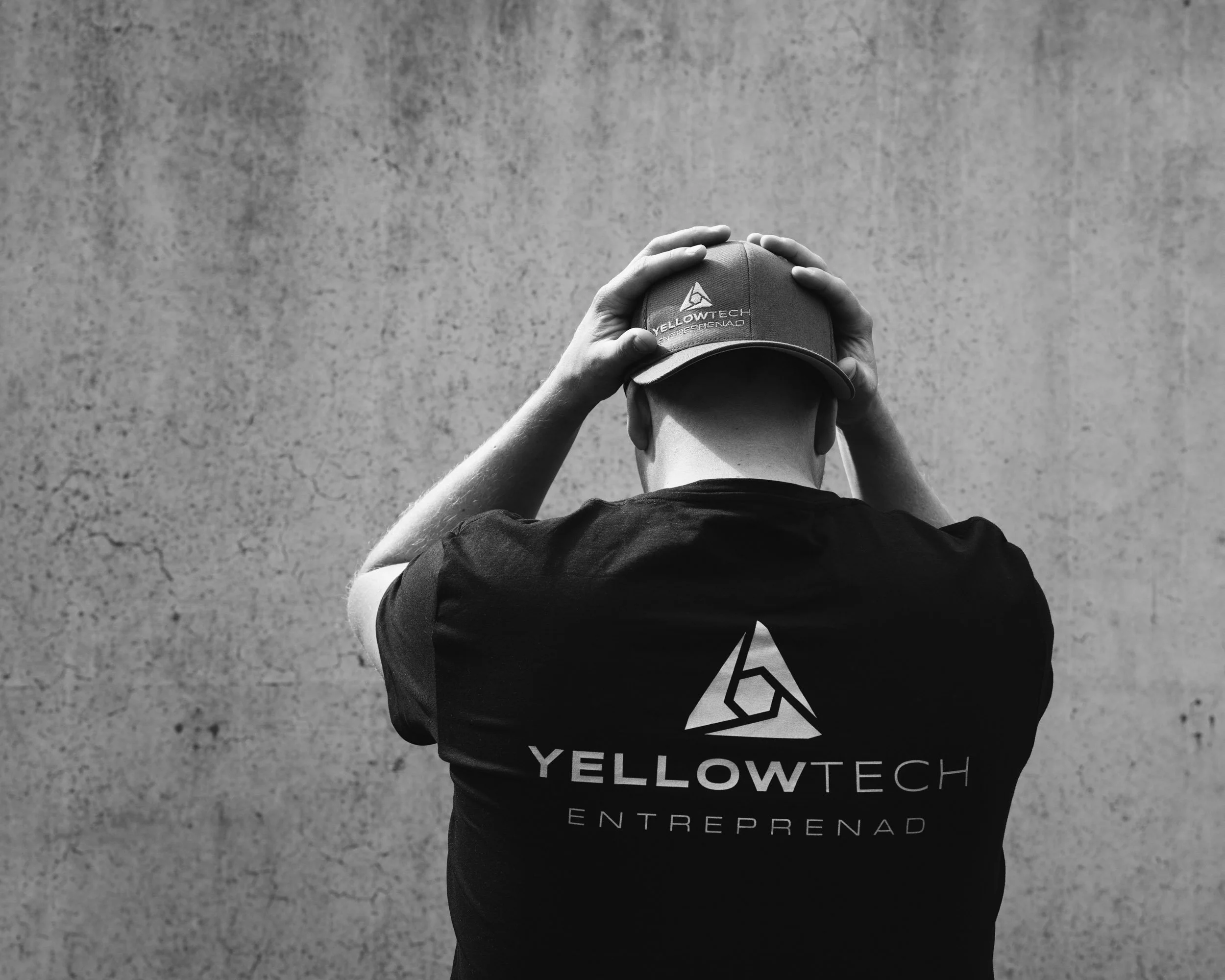 A person wearing a baseball cap and a t-shirt with the words 'YELLOWTECH ENTREPRENEUR' printed on it, standing against a textured wall, with their hands adjusting the cap.