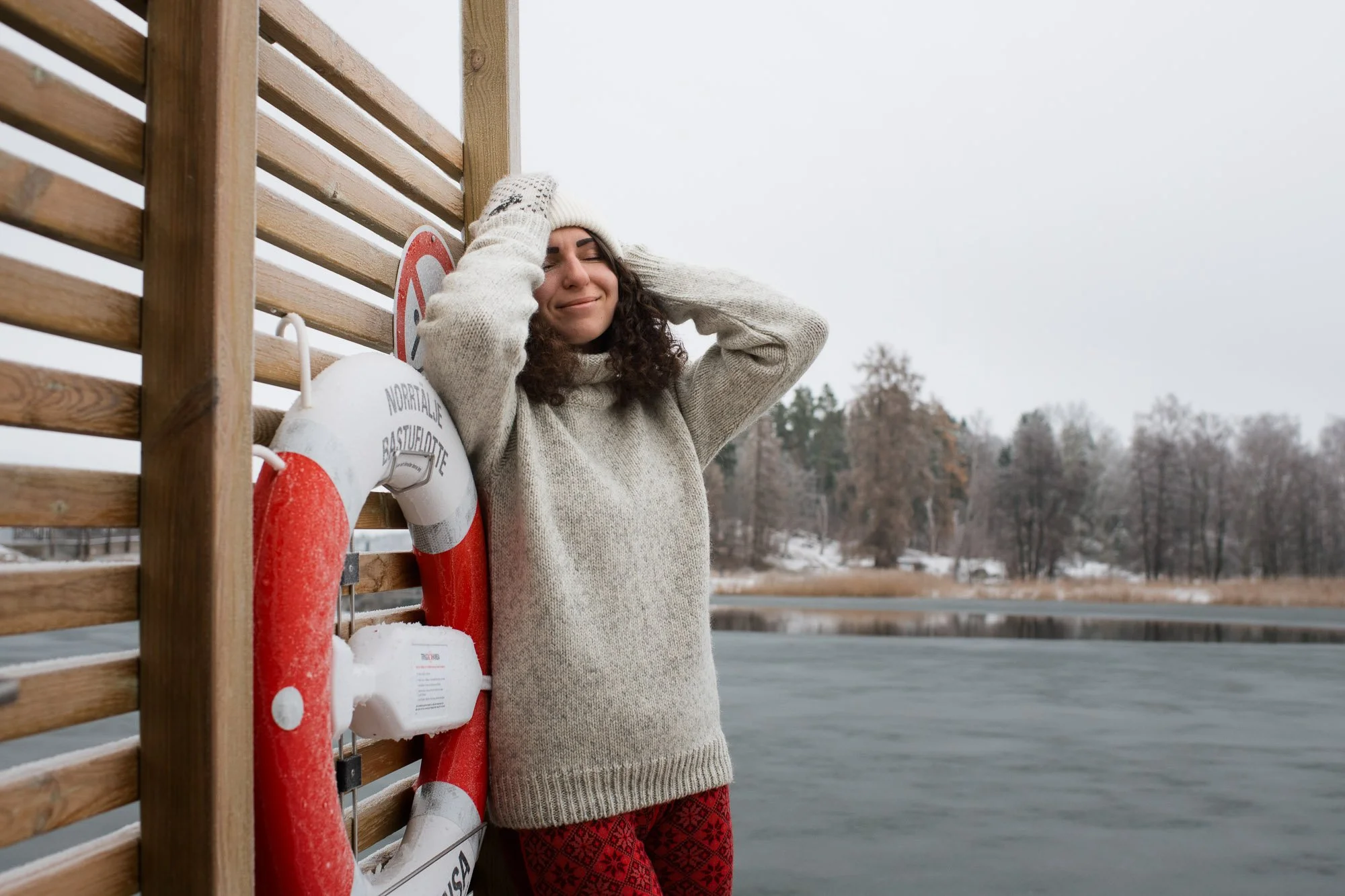 A woman in a winter sweater and patterned red pants standing next to a wooden fence with a life preserver attached, on a cold, overcast day near a body of water with trees in the background.