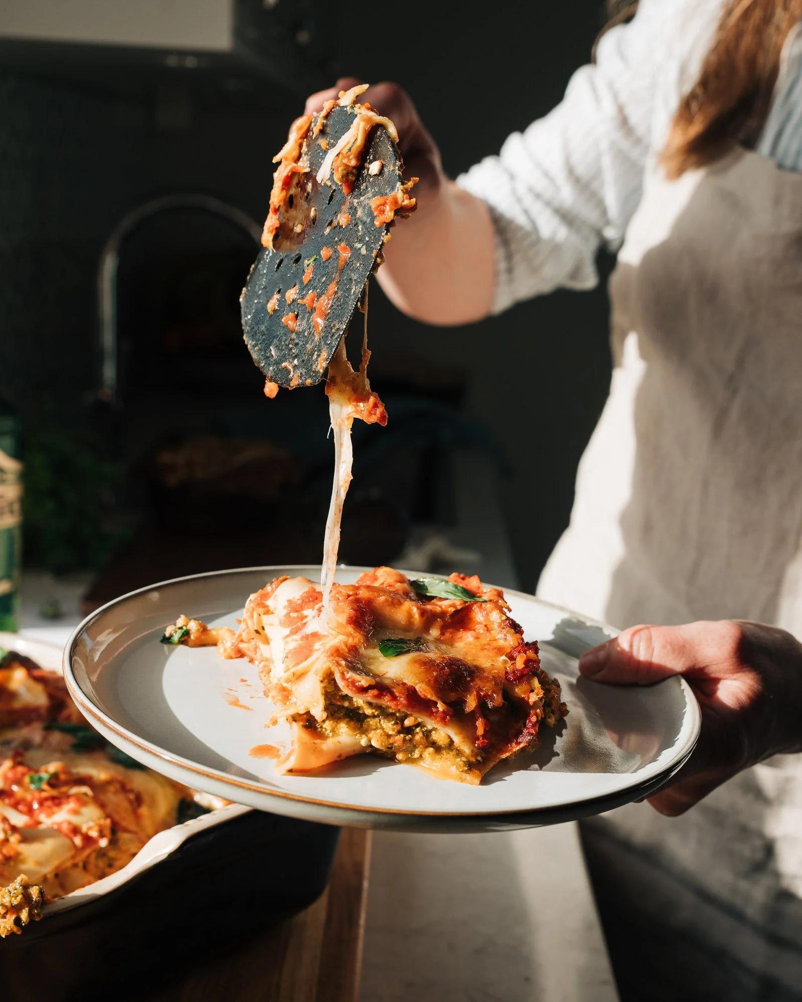Person serving a slice of layered lasagna with cheese and sauce onto a white plate.