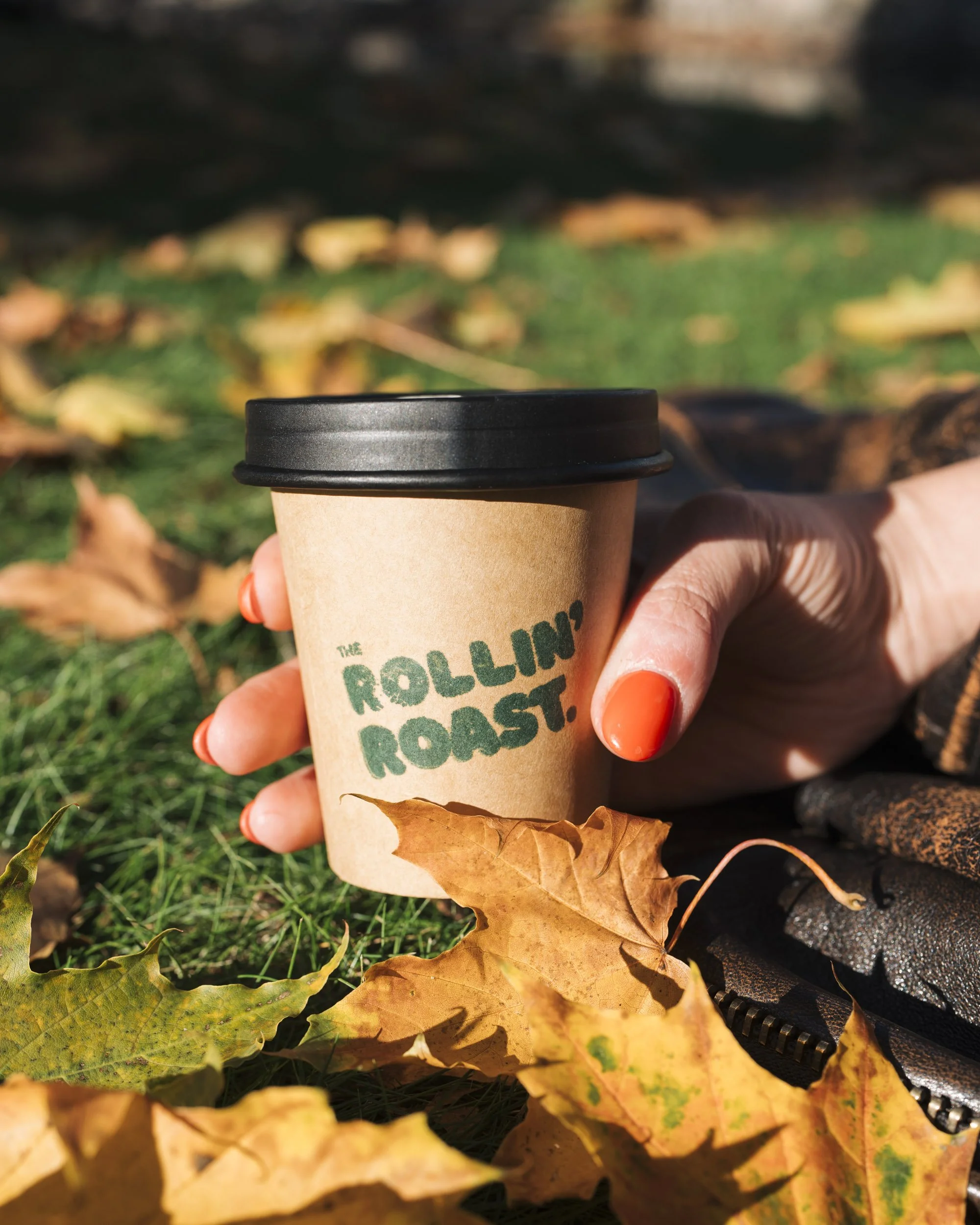 Person holding a disposable coffee cup with a black lid outdoors amid fallen fall leaves and grass.