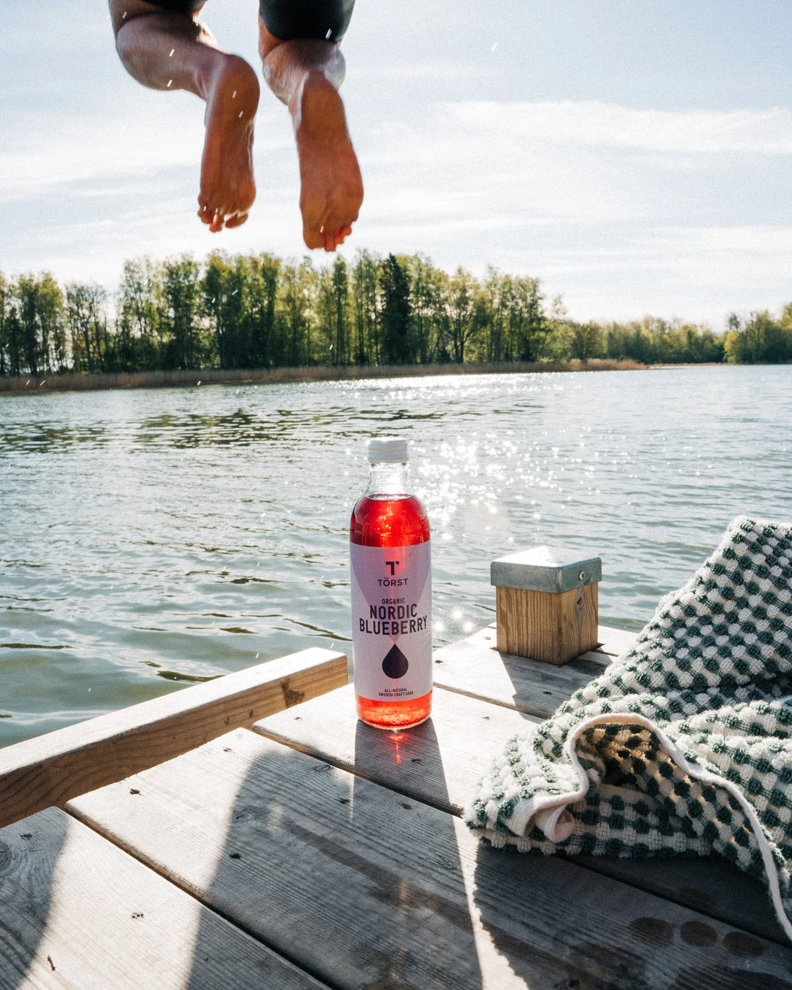A bottle of Nordic Blueberry soda on a wooden dock by a lake, with a person diving into the water in the background and a folded towel nearby.