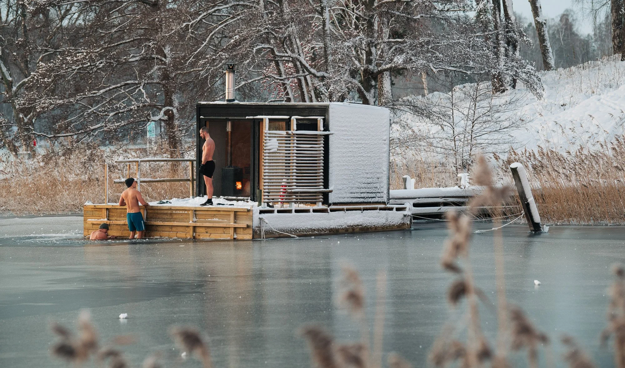 People swimming in an outdoor ice-covered lake near a snow-covered sauna in winter.