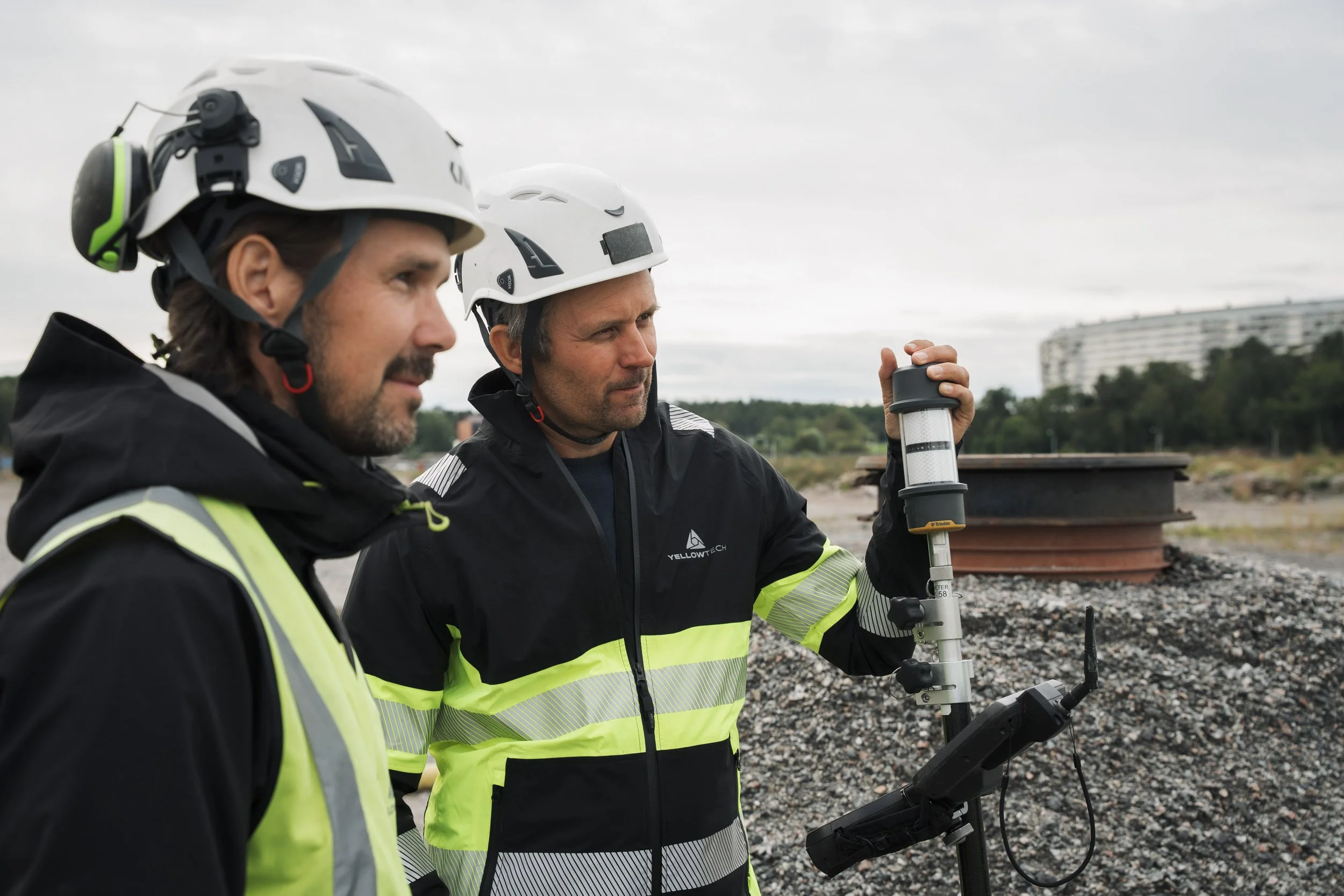 Two men wearing safety helmets and reflective jackets are working outdoors at a construction site. One is holding a surveying instrument.