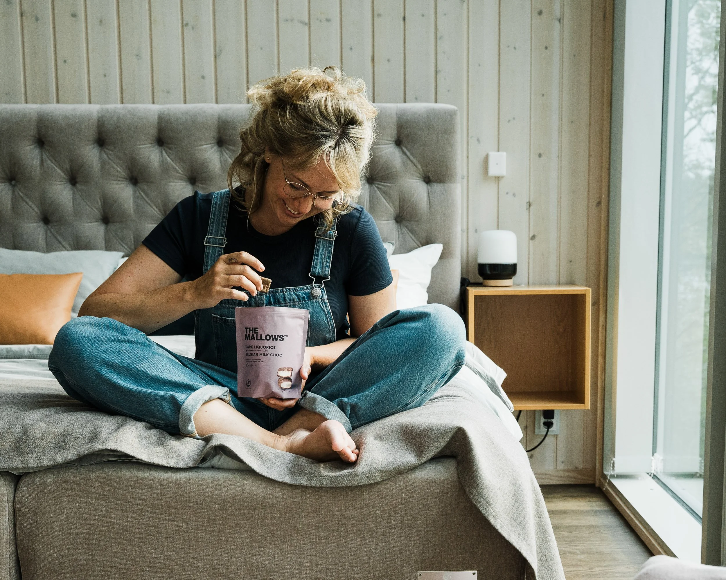 A woman with curly blonde hair, glasses, wearing a black shirt and denim overalls, sitting cross-legged on a bed, holding a packet labeled 'The Mallows,' smiling and looking down inside the packet.