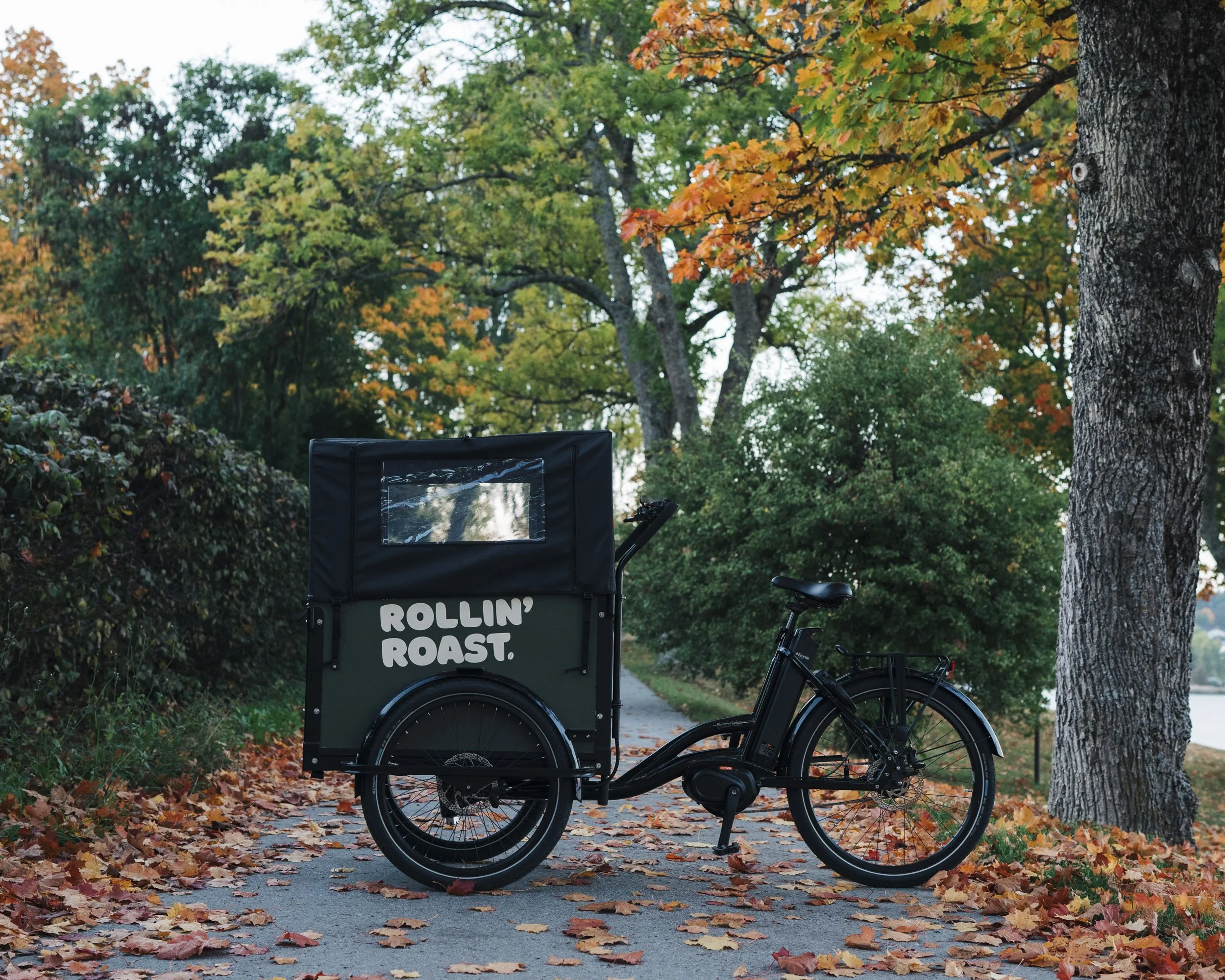 A black bicycle with a food cart attached, displaying the phrase "Rollin' Roast." parked on a leaf-covered pathway in a park with trees showcasing autumn foliage.