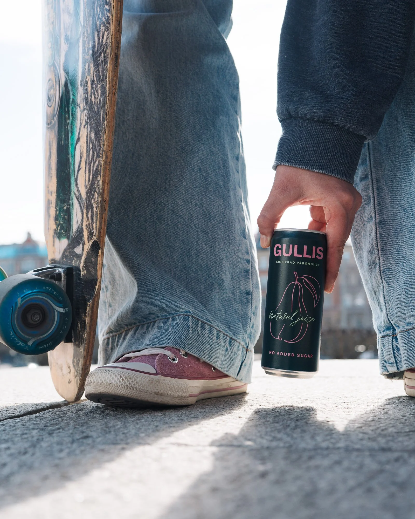 Close-up of a person wearing jeans and pink sneakers standing on the ground, holding a can of Gullis natural juice. A skateboard is resting nearby.