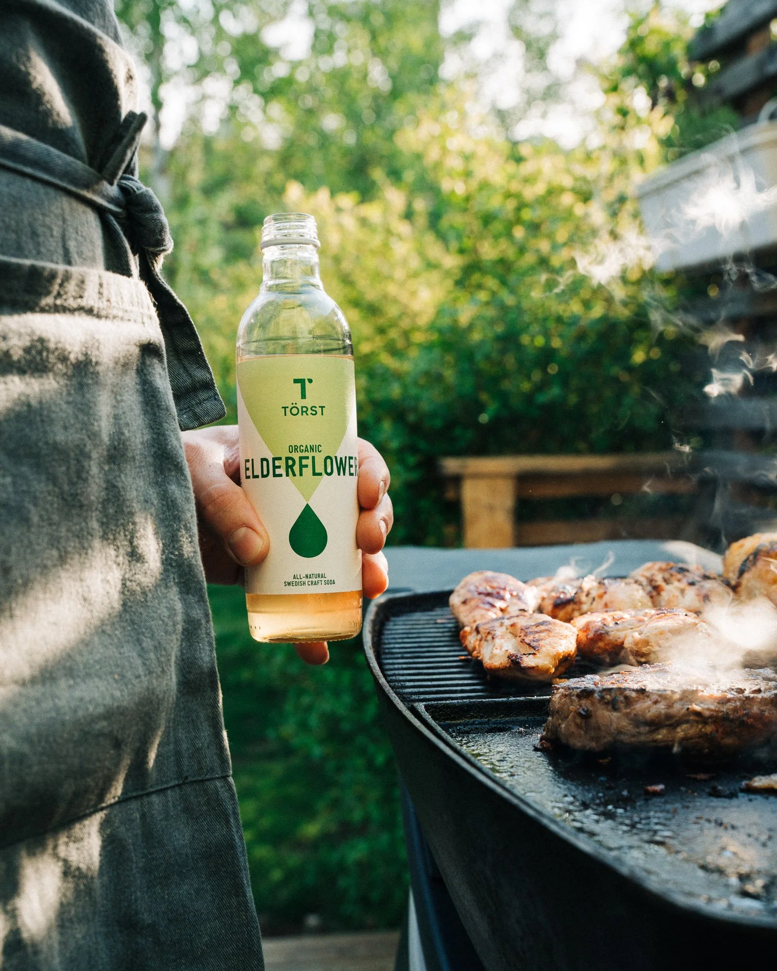 Person grilling chicken on a barbecue grill outdoors, holding a bottle of Törst Elderflower soda.