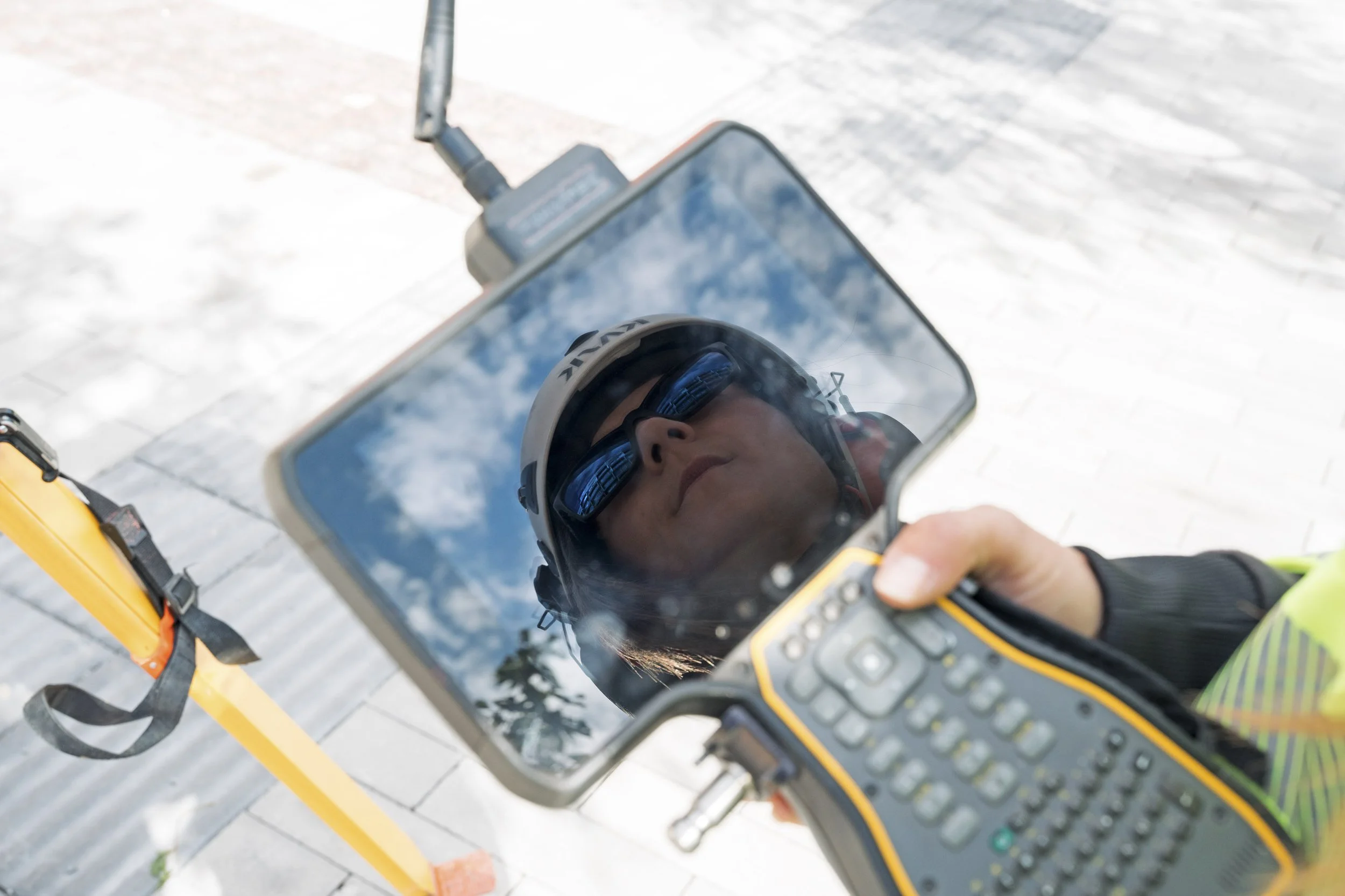 Male worker wearing a safety helmet and sunglasses reflected in a mirror attached to a surveying device at a construction site.