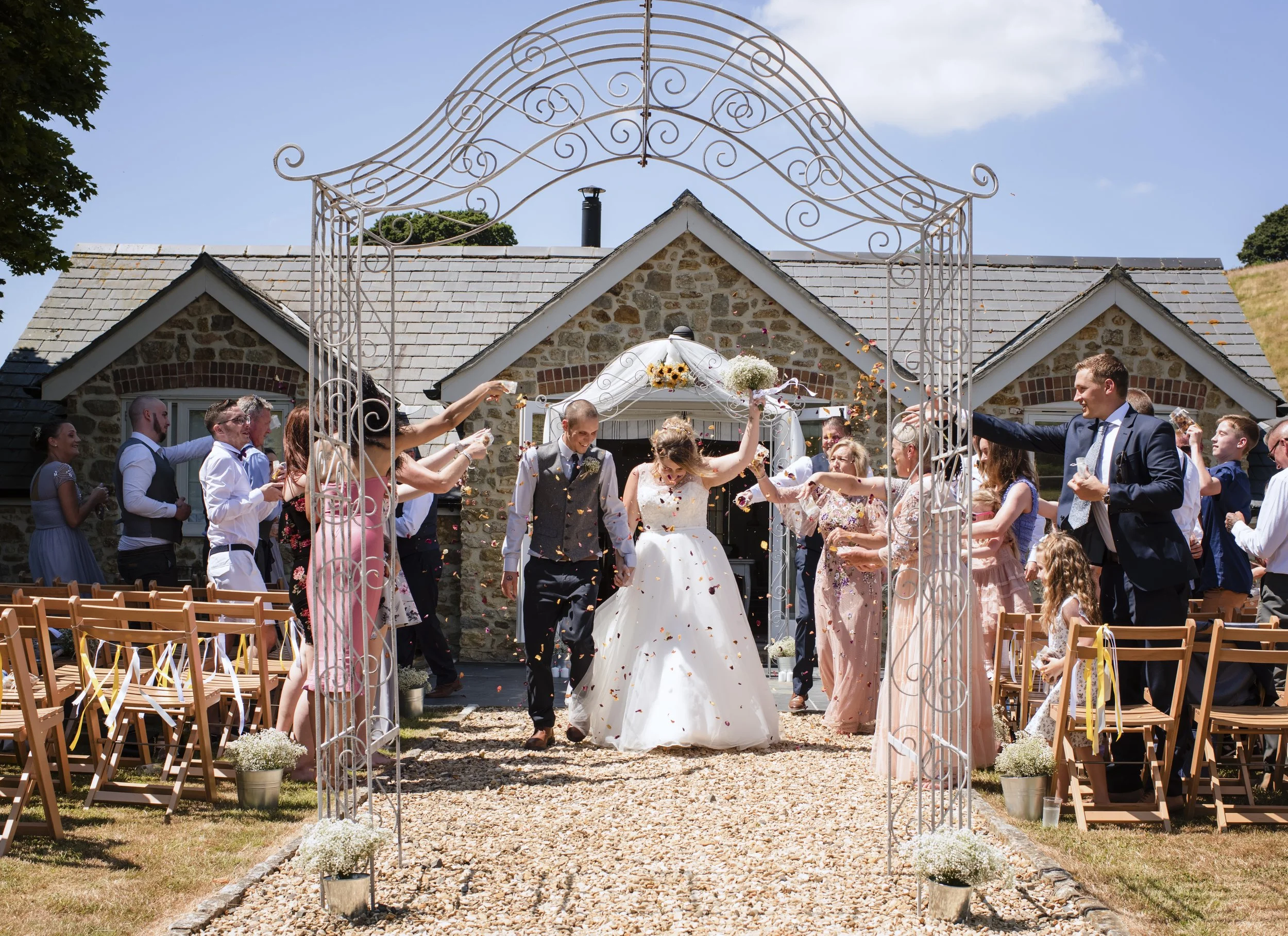 A wedding celebration outdoors with guests throwing flower petals as the bride and groom walk under a decorative metal archway, set against a stone house with grey shingled roof and a blue sky.