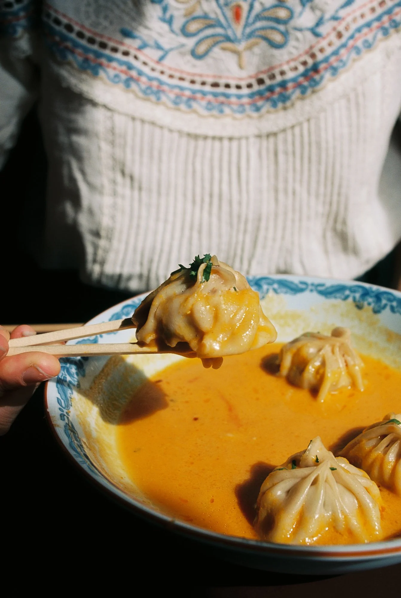 Close-up of a steamed dumpling held with chopsticks over a bowl of orange-colored dipping sauce, with more dumplings inside the bowl.