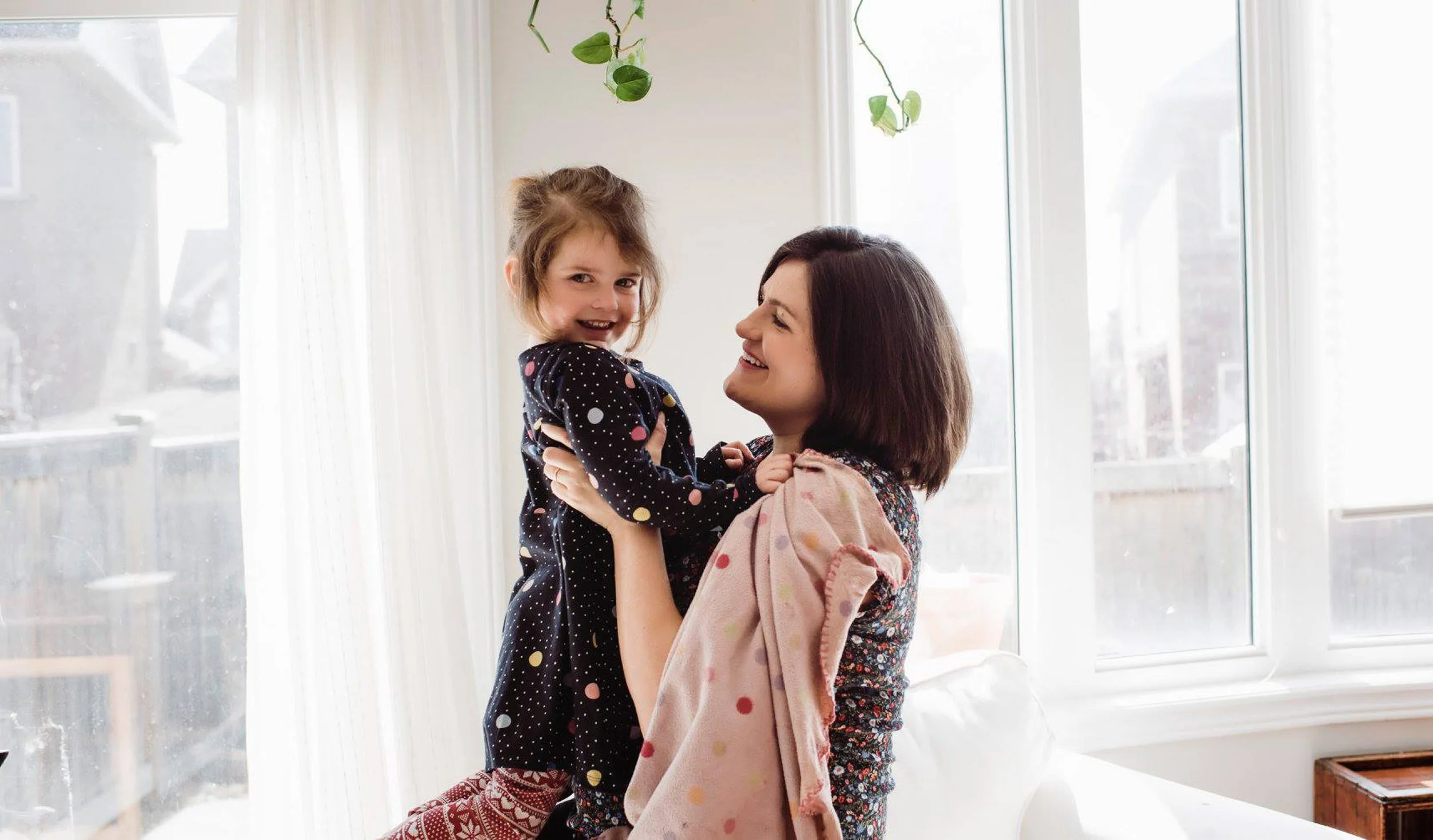 A woman with dark hair holding a smiling young girl with brown hair in a top knot, both standing near a large window with white curtains, with a hanging plant above them.