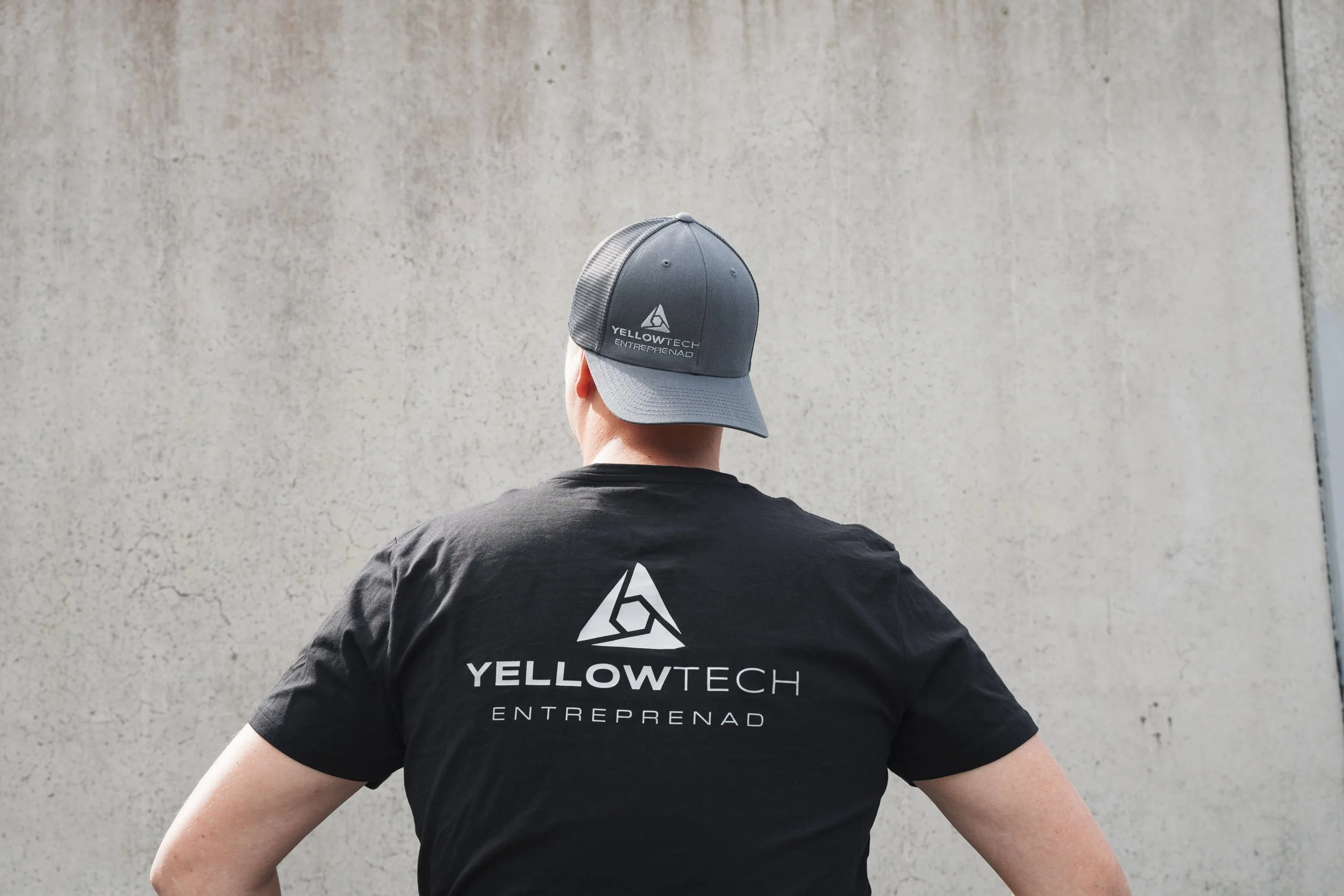 Person wearing a gray and black YELLOWTECH cap and a black T-shirt with the YELLOWTECH logo, standing in front of a plain concrete wall.