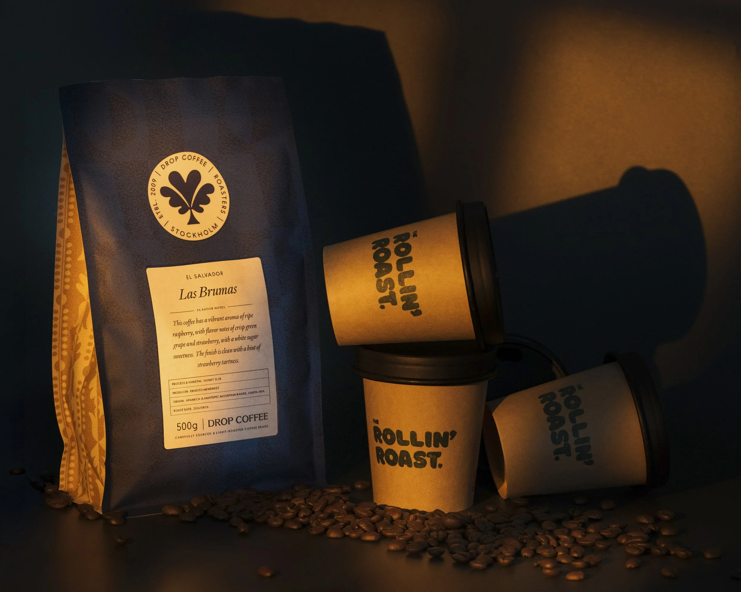 Coffee beans scattered on a surface with a package of coffee and a paper cup of coffee in front of a dark background with warm lighting.
