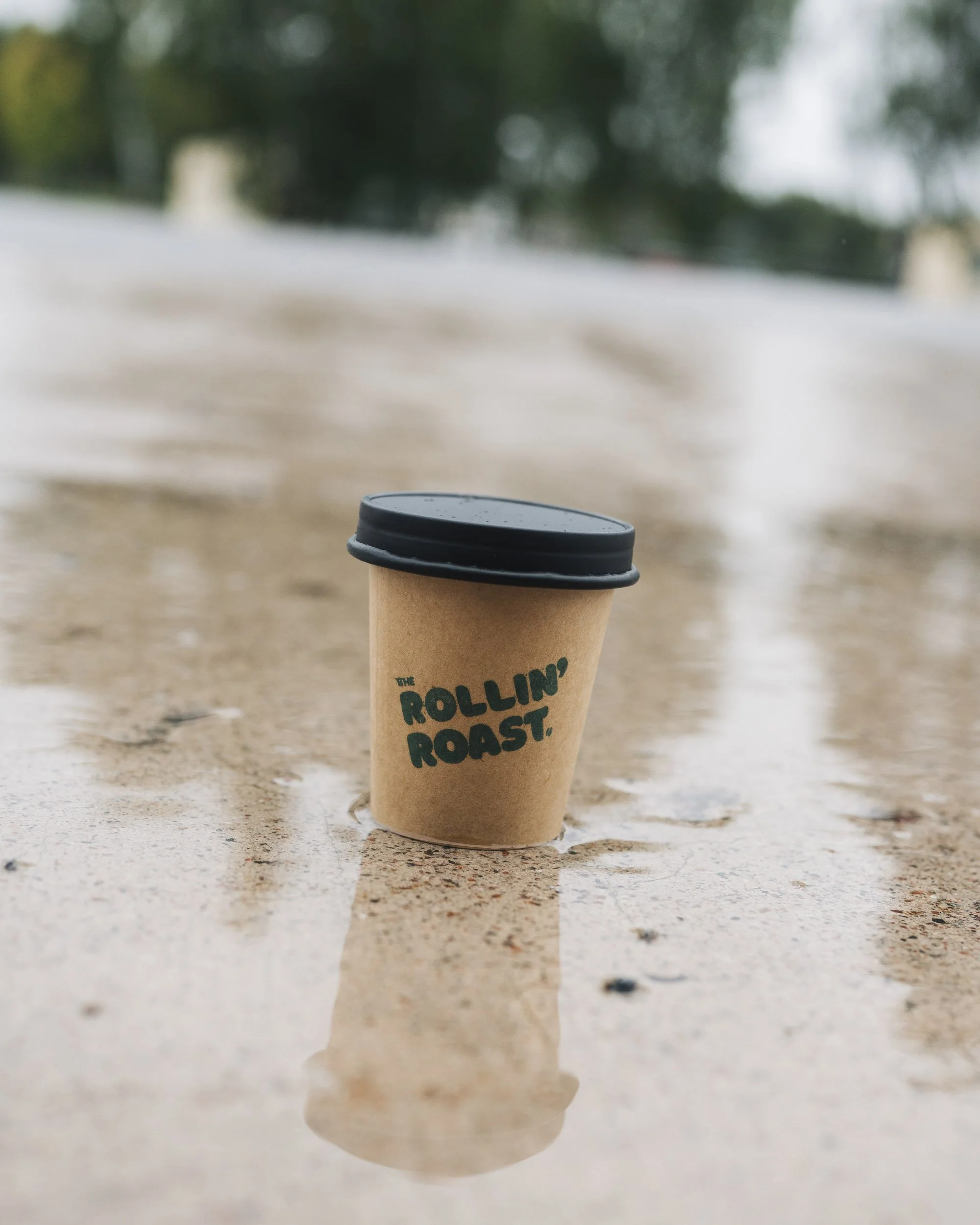 Close-up of a paper coffee cup with a black plastic lid, sitting on a textured surface, with a blurred outdoor background of trees and sky.