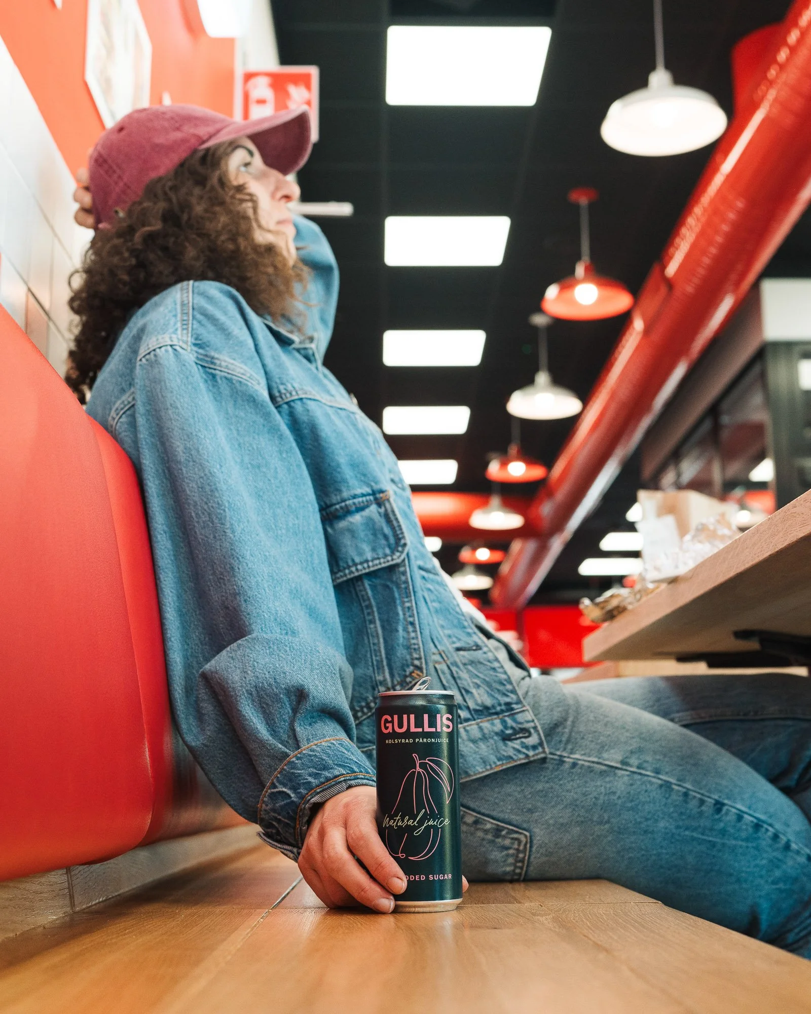 A woman with curly hair wearing a pink cap and denim jacket is sitting at a wooden table in a cafe, holding a can of Gullis natural juice with her right hand. The background features red and black ceiling lights and a red wall with fire safety signs.