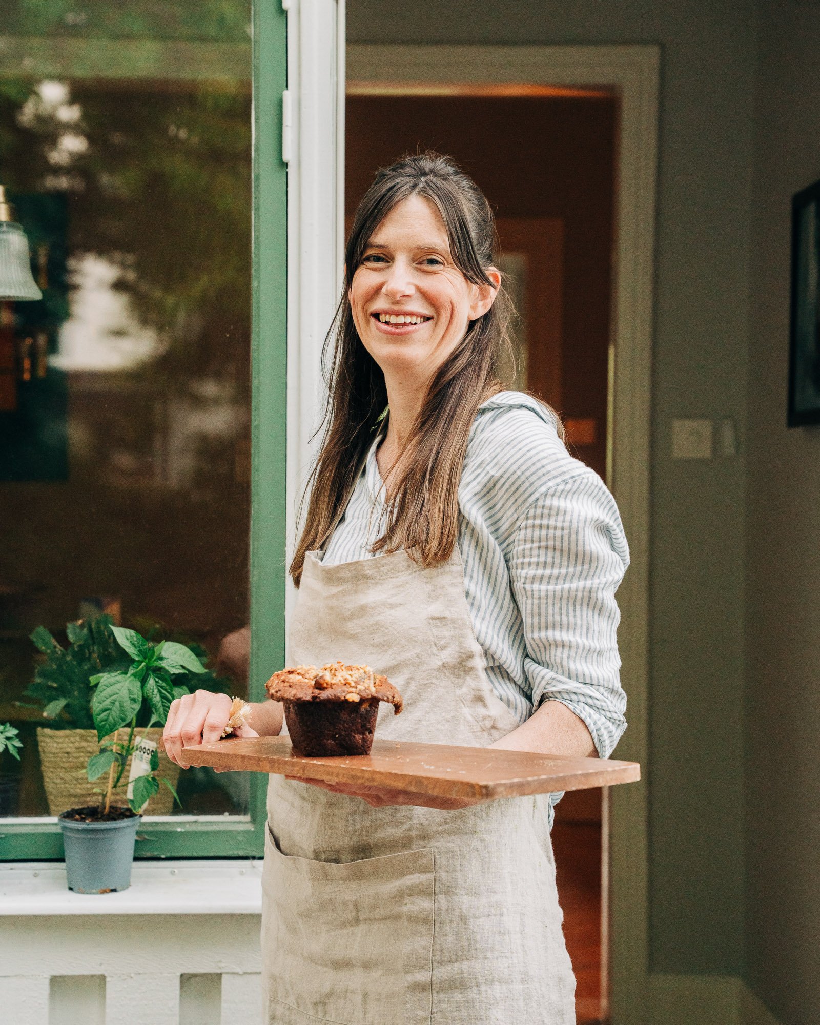 A smiling woman in a striped shirt and apron holding a wooden tray with a large cupcake, standing indoors near a green door with potted plants nearby.