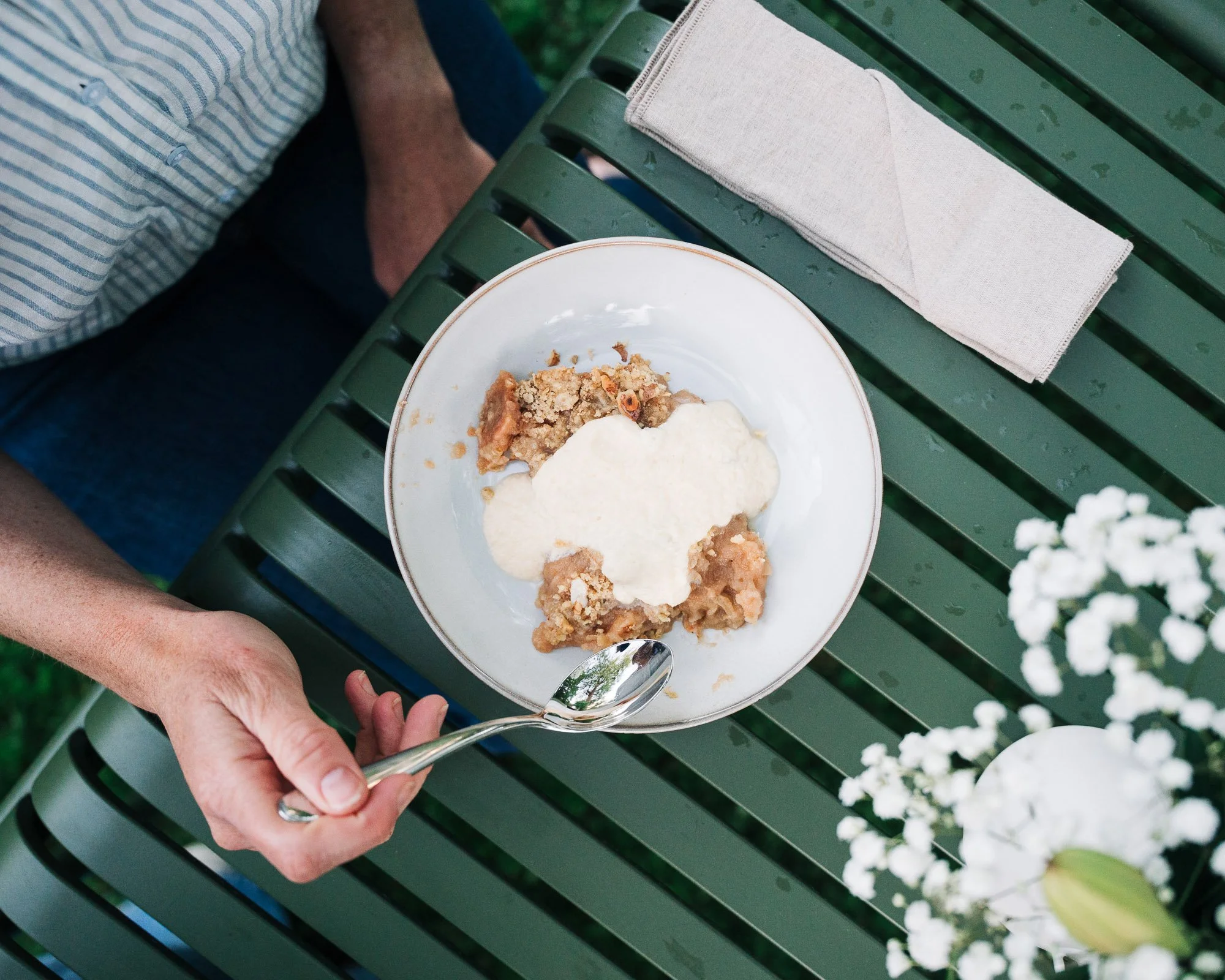 A person is holding a white bowl with a pie, whipped cream, and crumbs, on a green park bench with a beige cloth napkin and a spoon, with white flowers nearby.