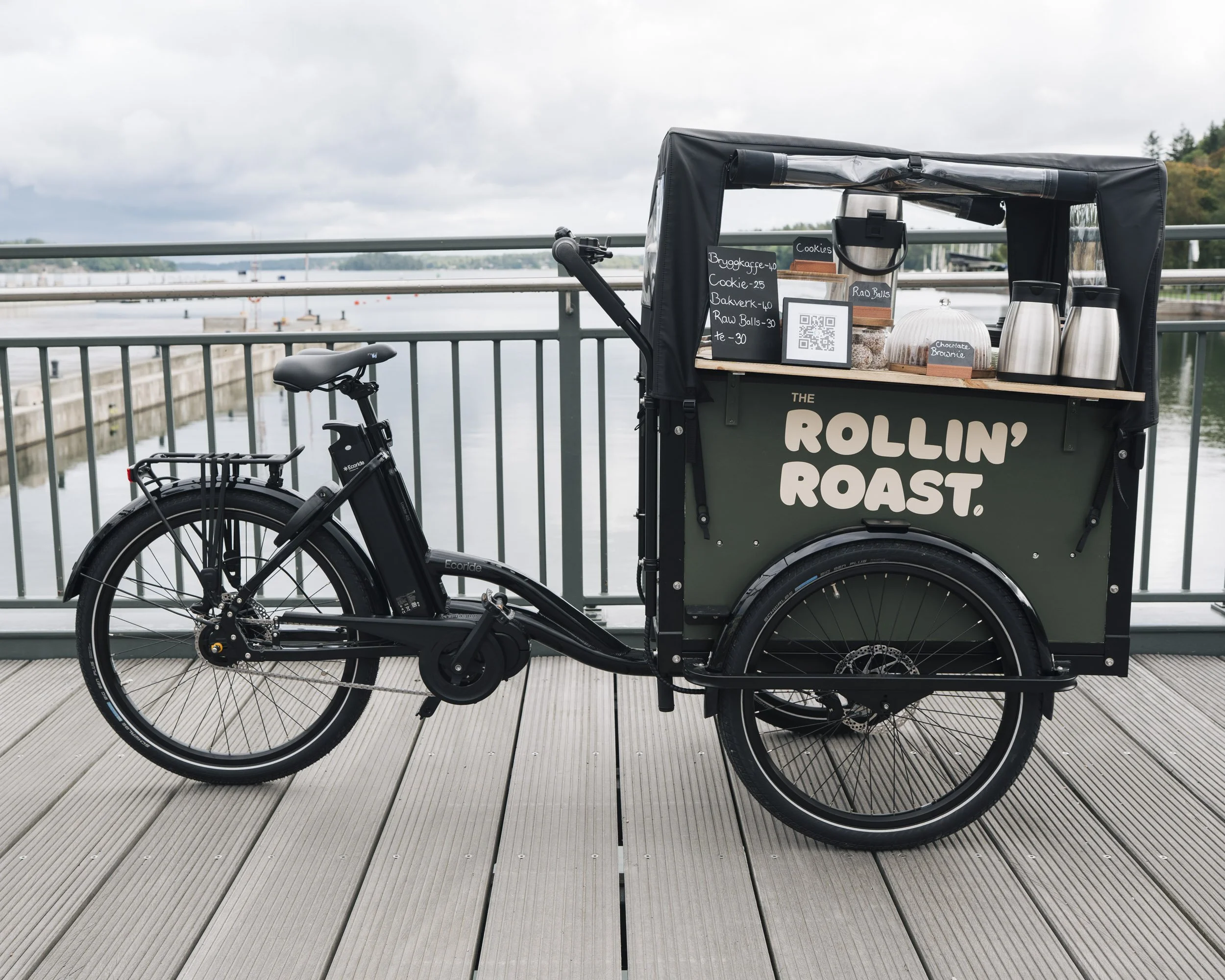 A bicycle with a food cart attached is parked on a wooden boardwalk near a body of water, with a metal railing and a cloudy sky in the background.