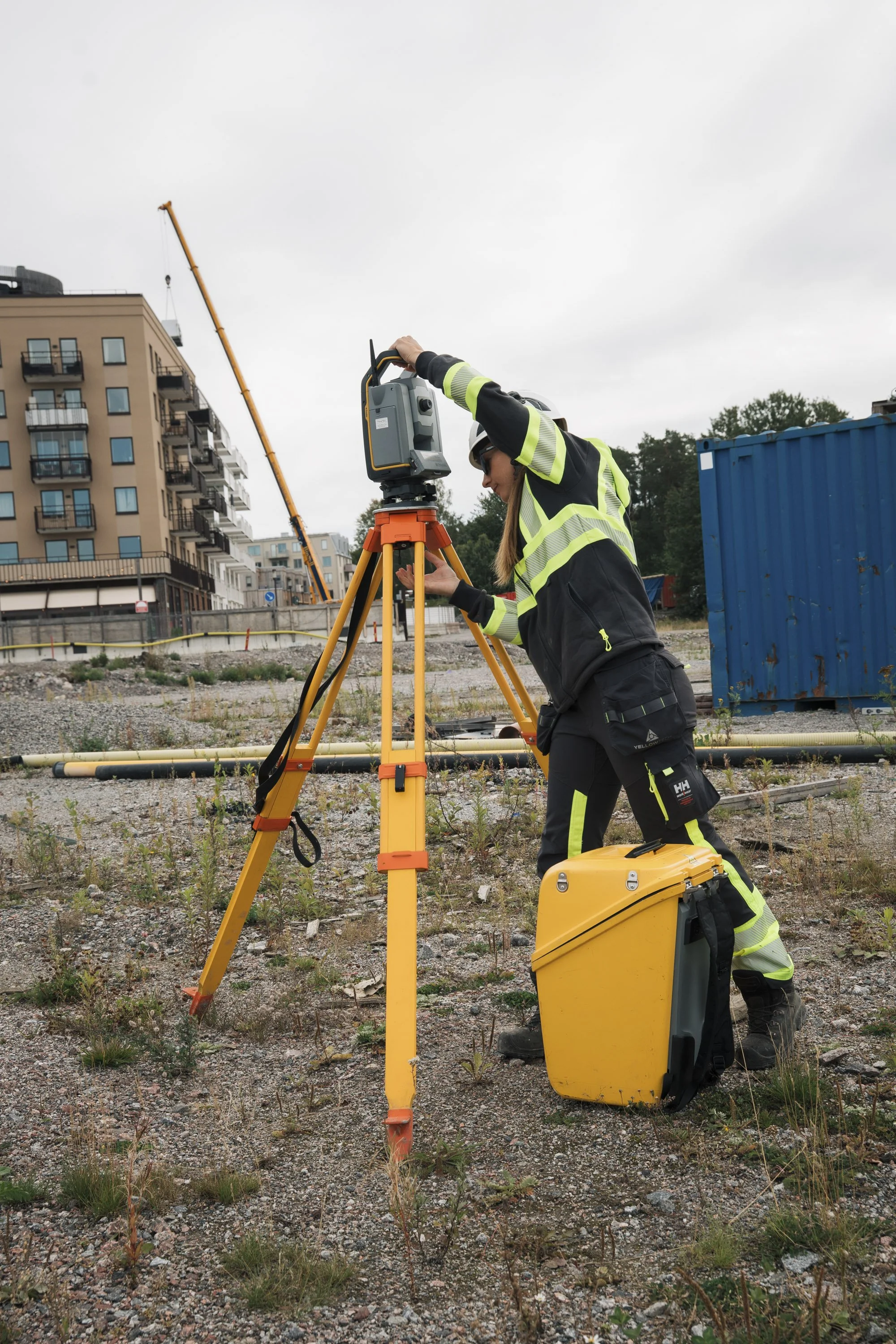 A construction worker at a construction site, wearing a high-visibility jacket and helmet, uses surveying equipment on a tripod. There are buildings and construction cranes in the background.