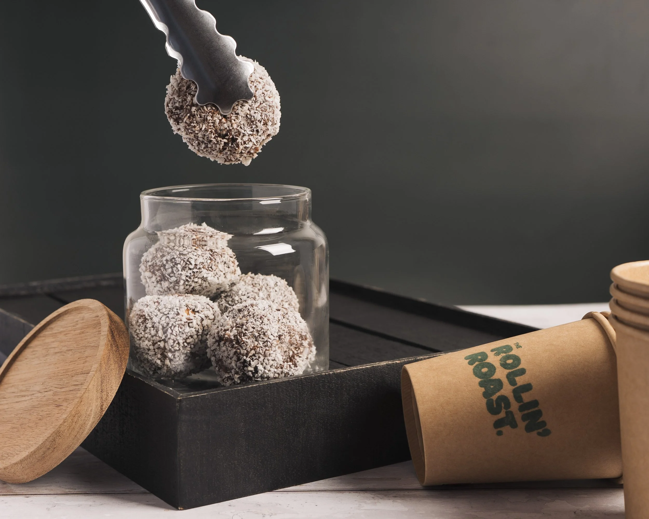 A stack of round, chocolate truffles covered in shredded coconut, with one being held up by tongs over a glass jar on a black surface.