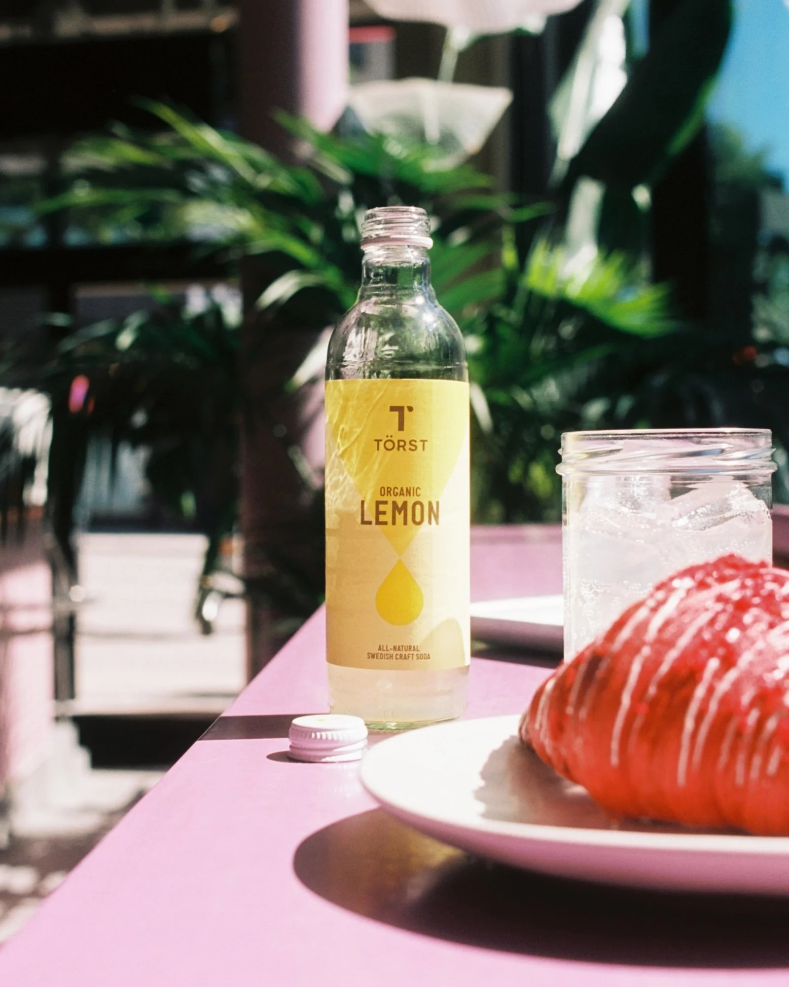 A clear glass bottle of organic lemon Swedish craft soda on a pink table, with a white bottle cap nearby and a red pastry on a white plate in the foreground. Blurred green plants and dark furniture are in the background.