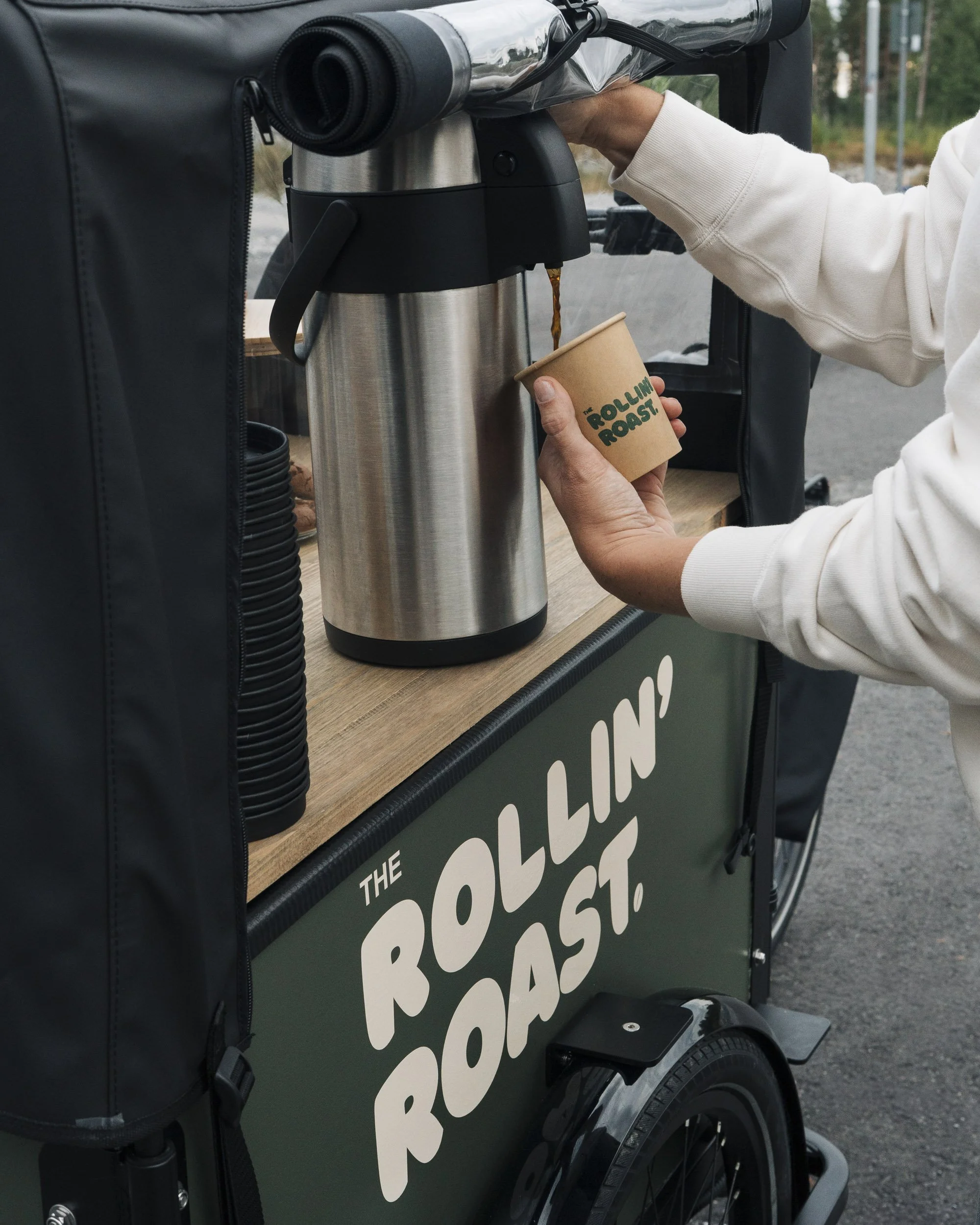 A black and green food truck with a wooden countertop, dispensing hot coffee from a large stainless steel coffee urn into a disposable paper cup held by a person wearing a white sweatshirt.