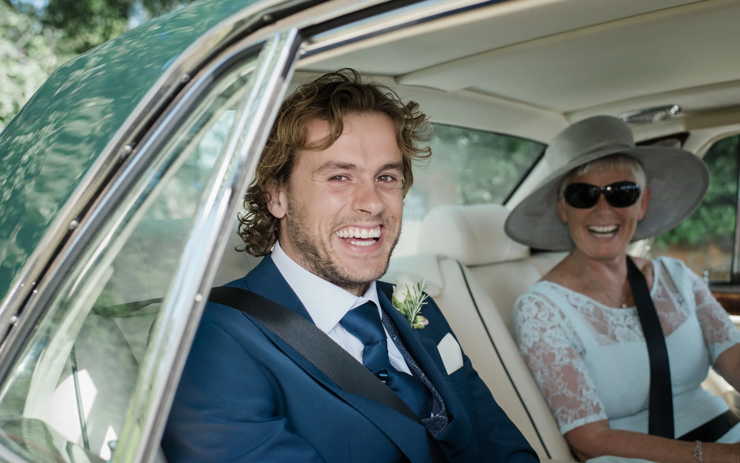 A young man in a blue suit and a boutonniere, smiling, sitting in a car with an older woman in a white dress and large sun hat, also smiling in the back seat.