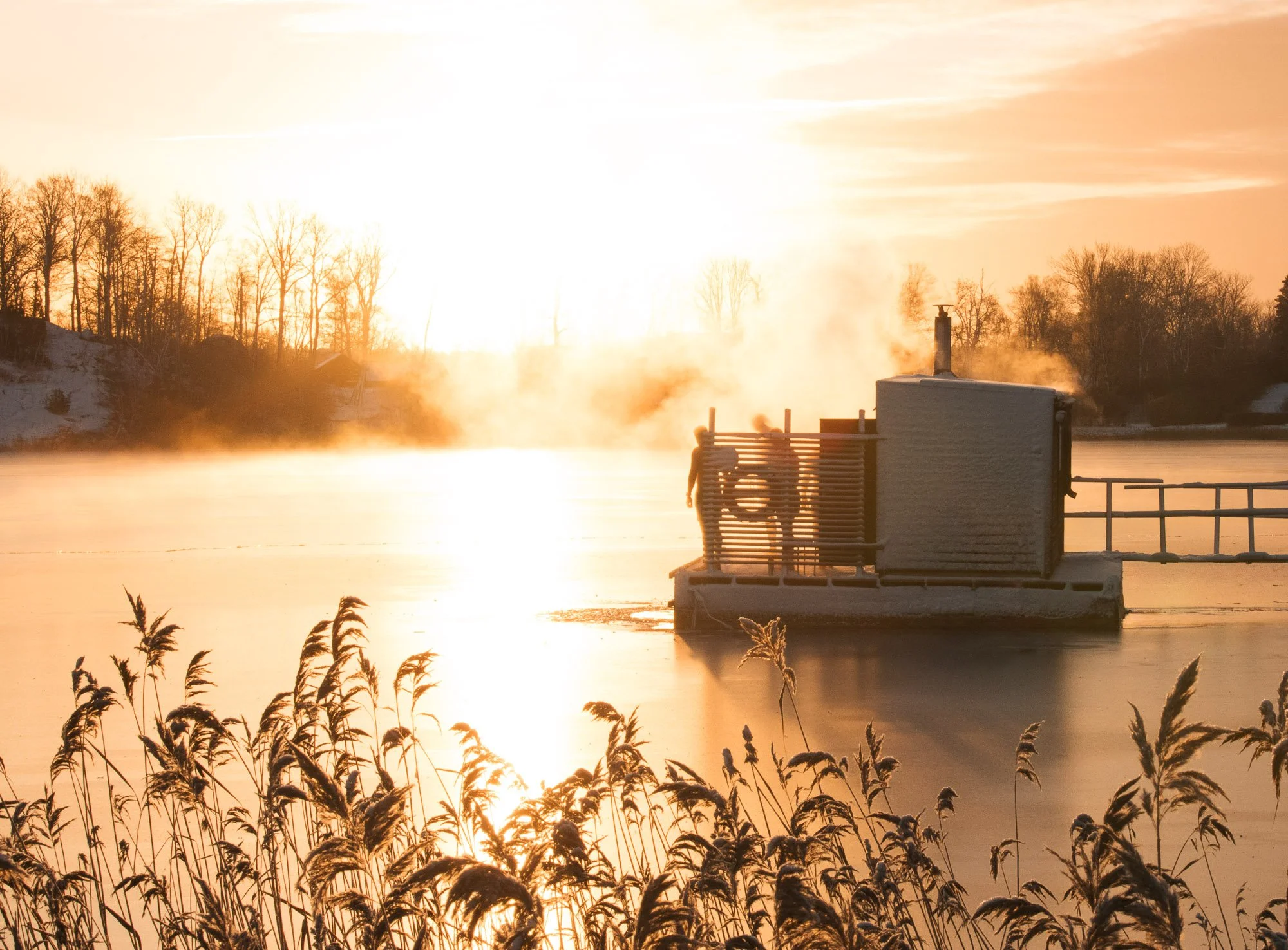 Sunset over a frozen lake with steam rising from the water, trees in the background, and a small building with a chimney on a floating dock.